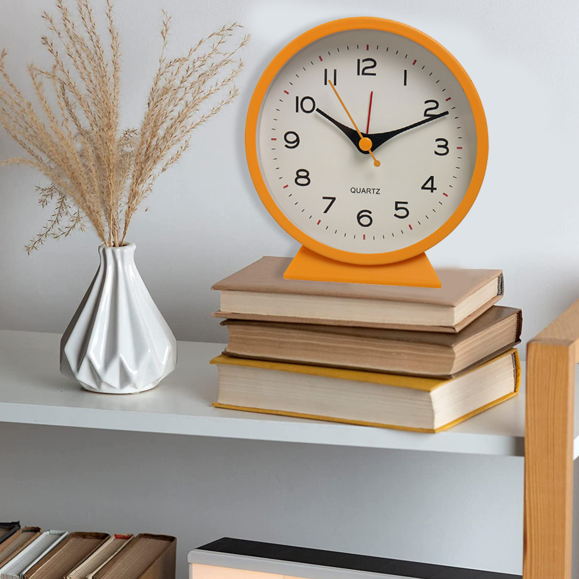 Eine orange Quarzuhr steht auf einem Stapel Bücher neben einer weißen Vase mit getrocknetem Gras.