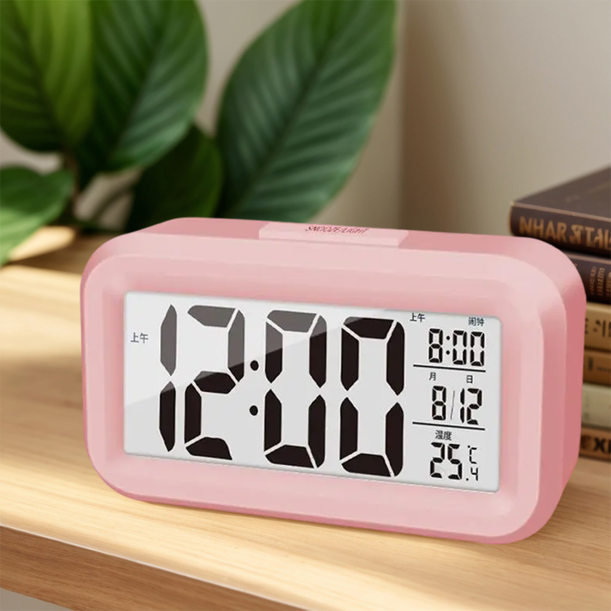 Ein rosafarbener Digitalwecker zeigt 12:00 an, neben Büchern und Pflanzen.