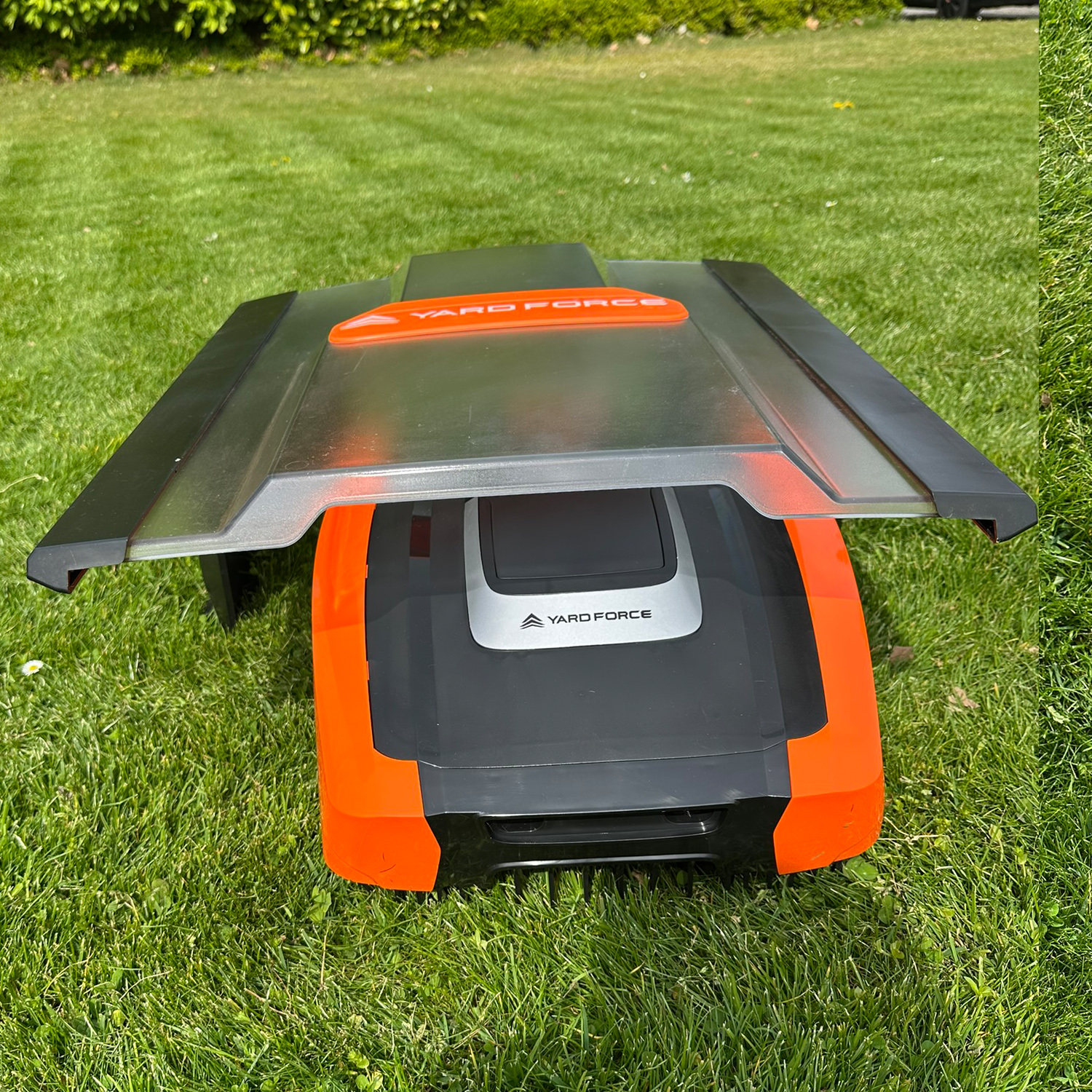 Ein orange-schwarzer Mähroboter mit offenem Deckel steht auf einem grünen Rasen.
