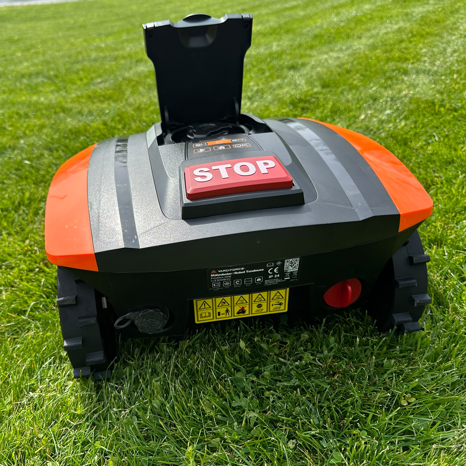 Ein Mähroboter mit orangen und grauen Akzenten steht auf einem grünen Rasen. Die Haube ist geöffnet.