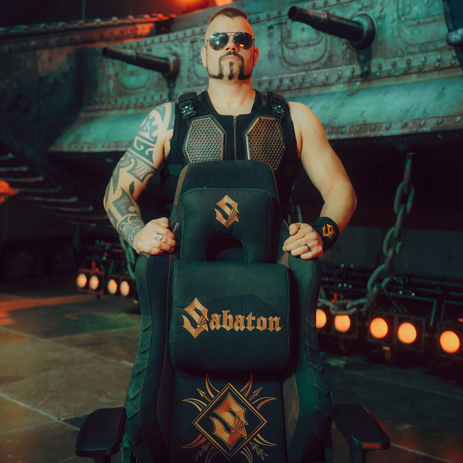 Mann in taktischer Ausrüstung, Sonnenbrille und Tattoos sitzt in einem Sabaton-Gaming-Stuhl.