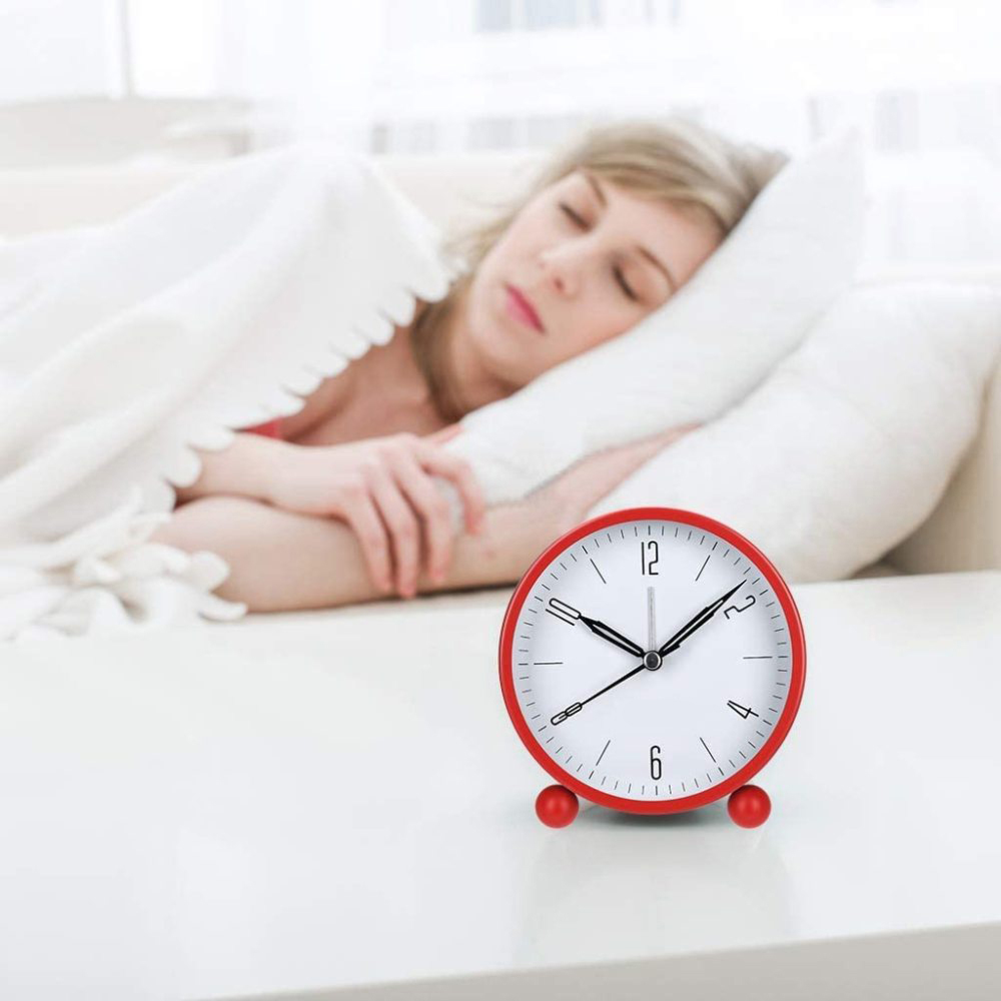 Eine Frau schläft unter einer Decke, ein roter Wecker steht auf dem Tisch.
