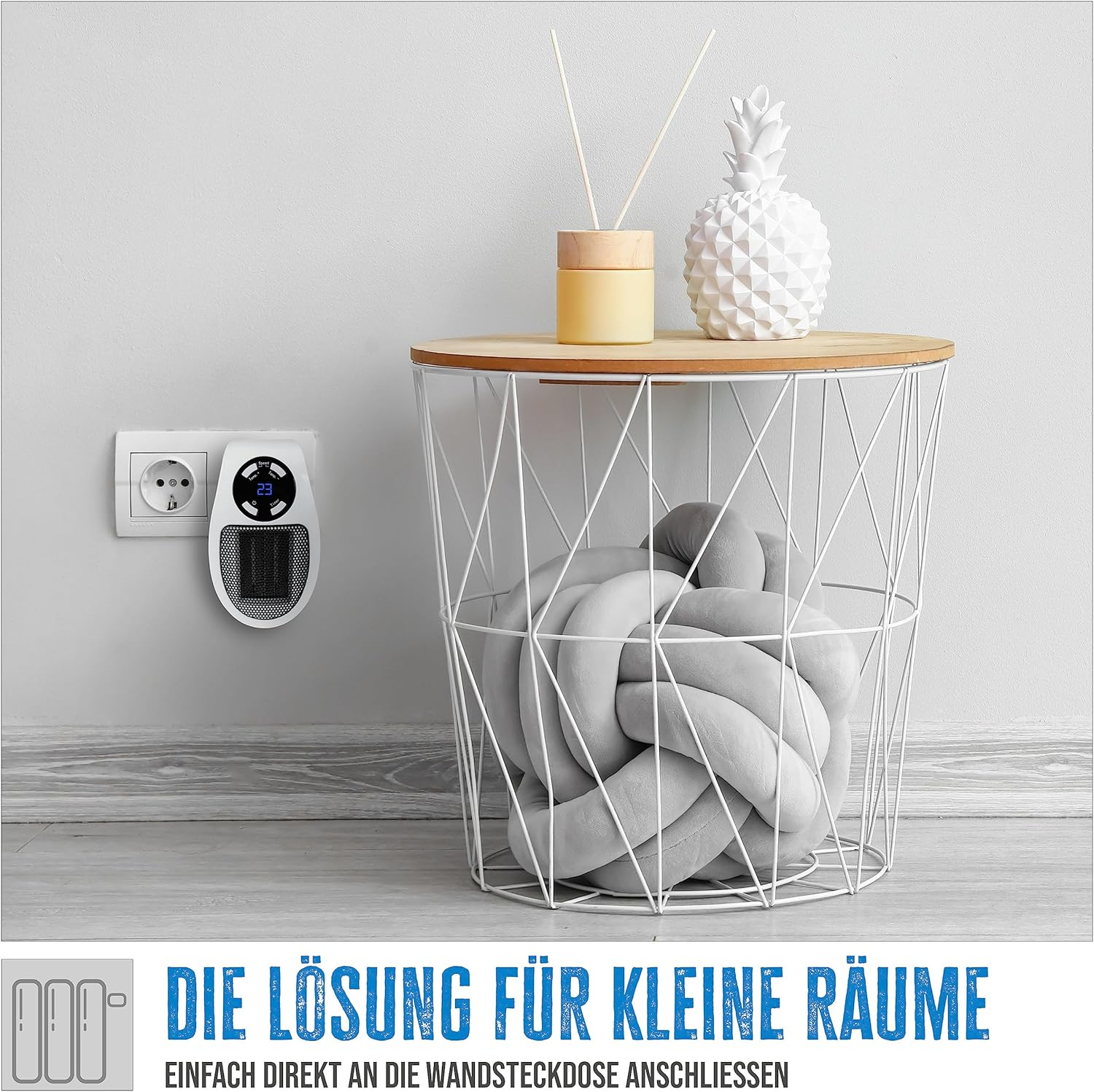 Ein weißer Raumheizer ist in eine Steckdose gesteckt. Daneben ein weißer Drahtkorb mit Kissen.