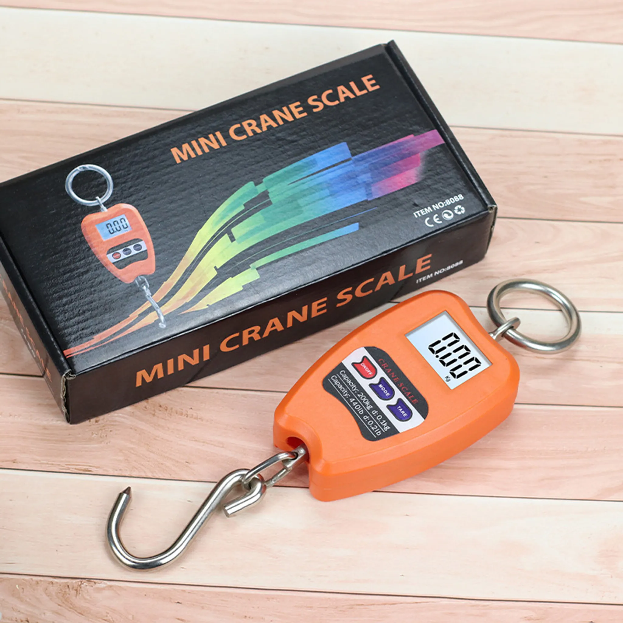 Eine orangefarbene Mini-Kranwaage liegt neben ihrer schwarzen Verpackung auf Holzhintergrund.