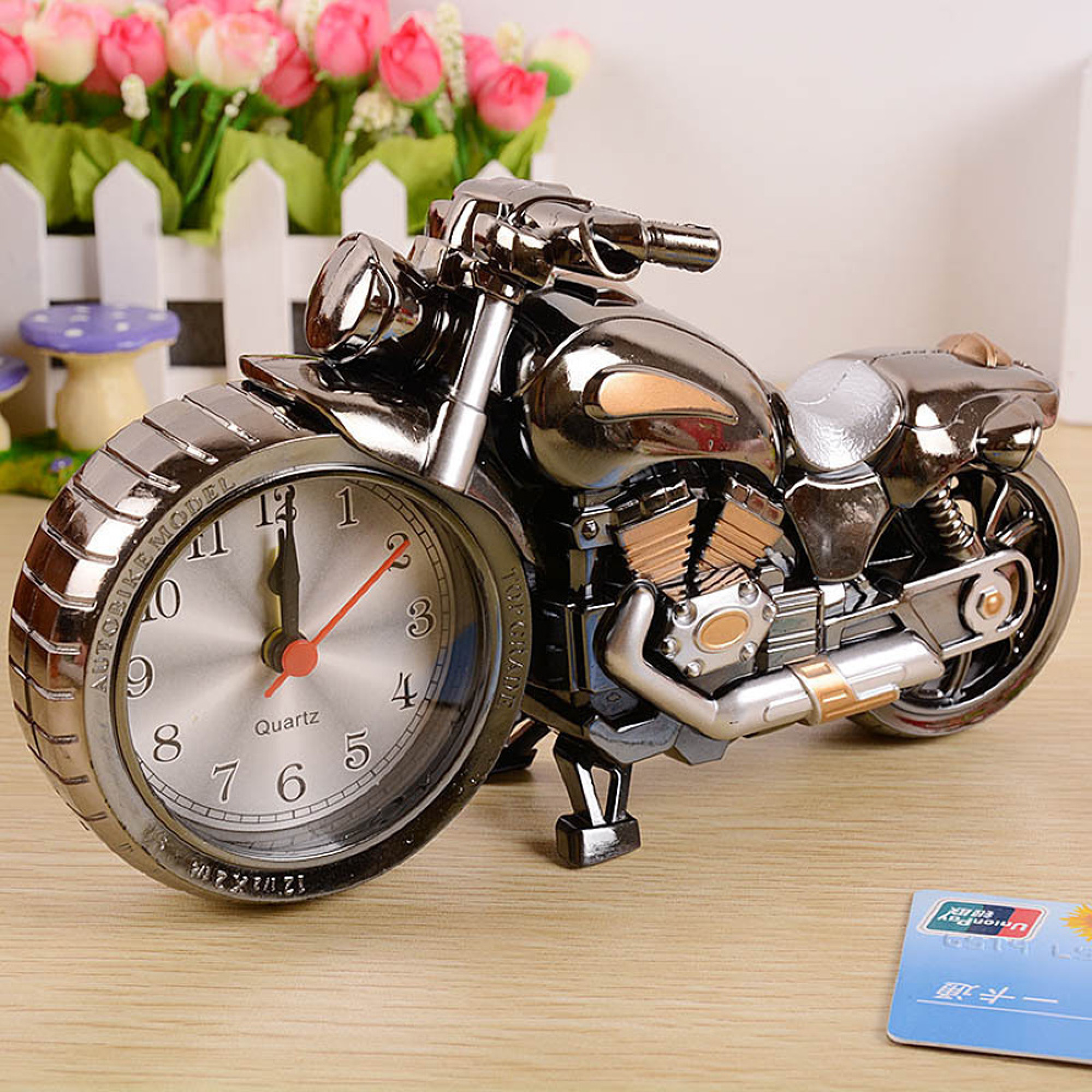 Ein silberner Motorrad-Wecker steht auf einem Holztisch. Blumen sind im Hintergrund.