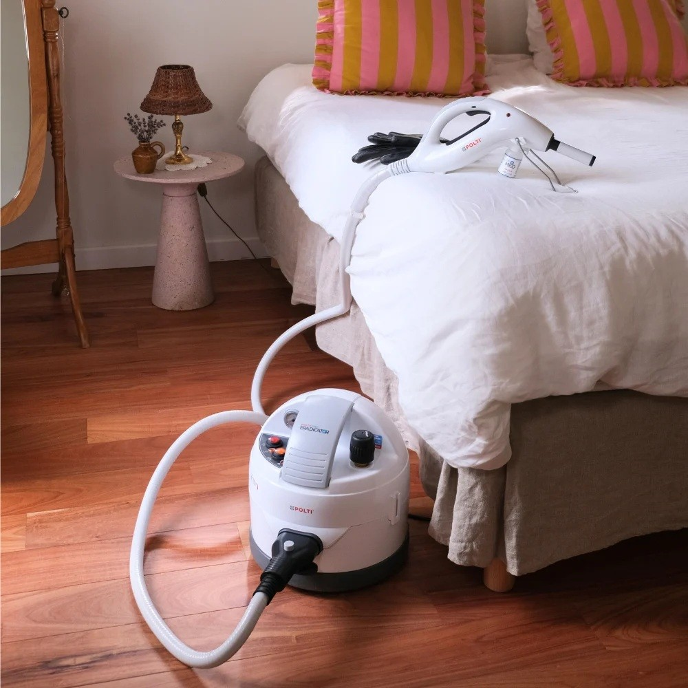 Ein weißer Dampfreiniger mit Schlauch liegt auf einem Bett. Ein Nachttisch mit Lampe steht daneben.