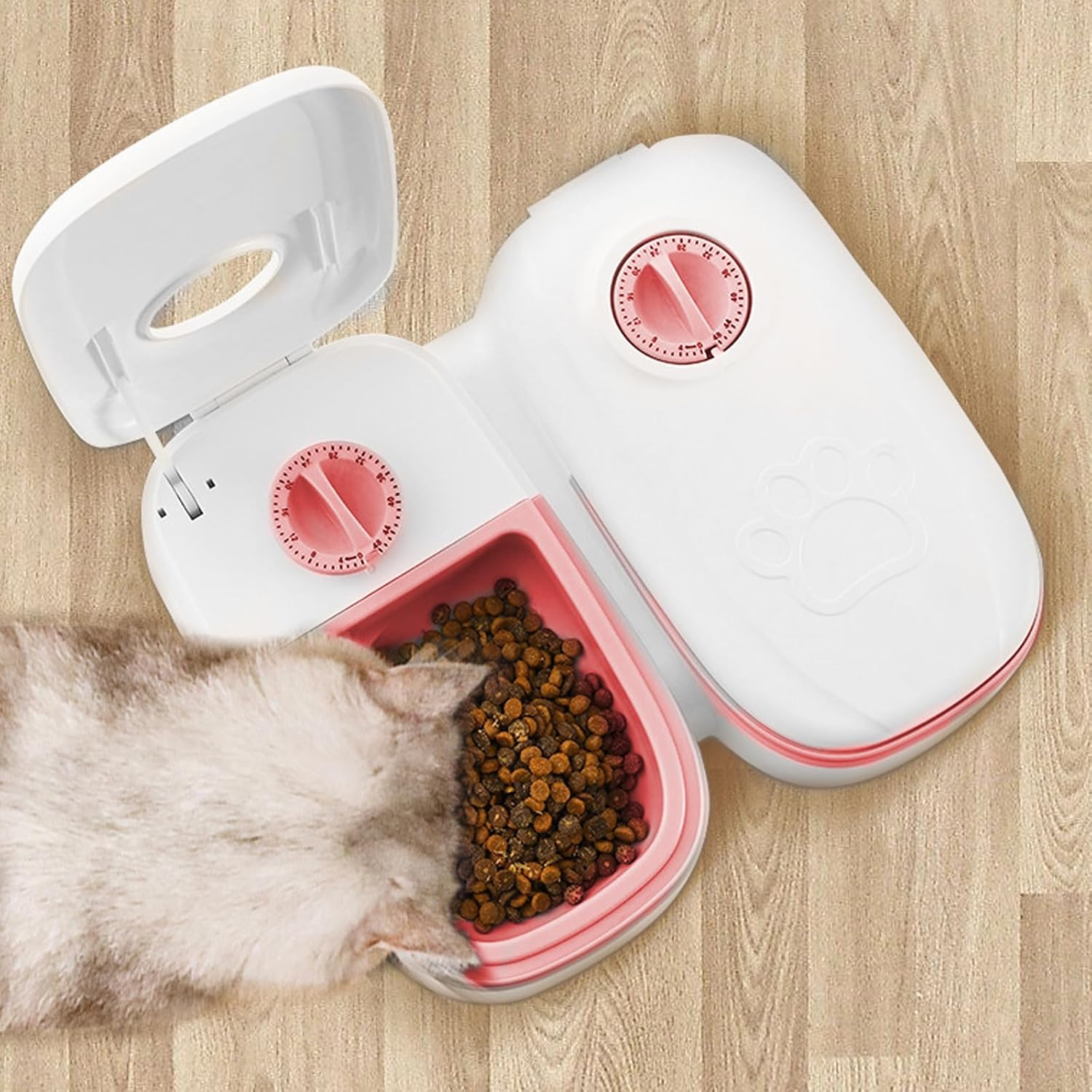 Eine Katze frisst aus einem weißen Futterspender mit rosa Schüssel auf Holzboden.