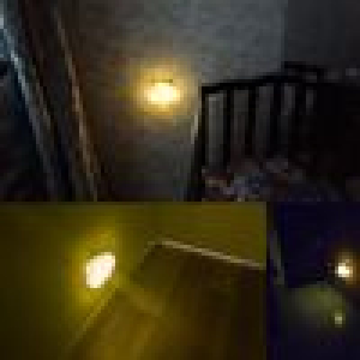 Eine Wandlampe leuchtet gelb im dunklen Flur neben einer Treppe mit Geländer.