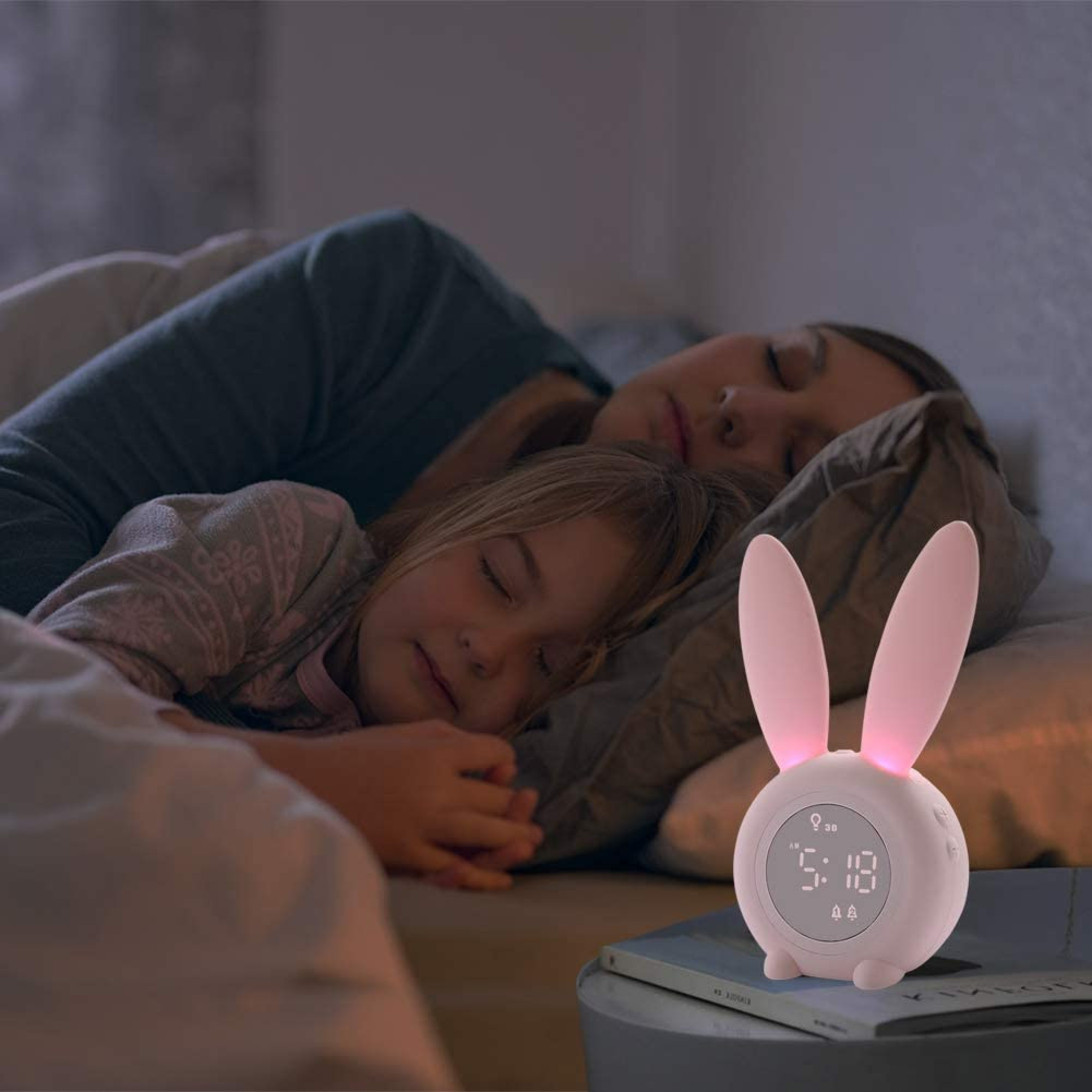 Ein rosafarbenes Hasenlicht mit digitaler Uhr steht neben einem schlafenden Kind und einer Frau.