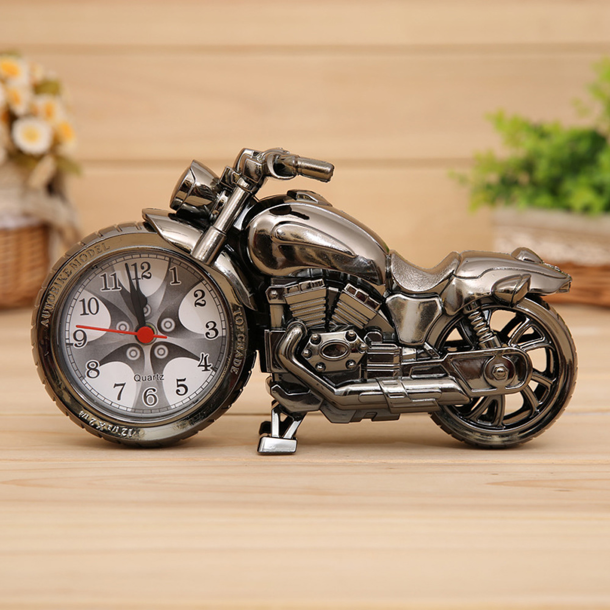 Ein silberner Motorrad-Wecker steht auf einer Holzoberfläche, im Hintergrund unscharfe Blumen und Grün.