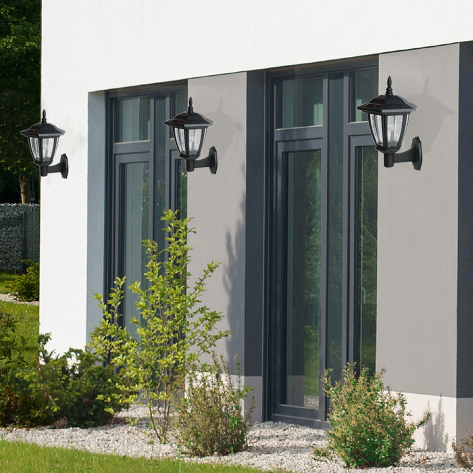 Drei schwarze Wandleuchten mit Solarmodul an einem Haus über Büschen.