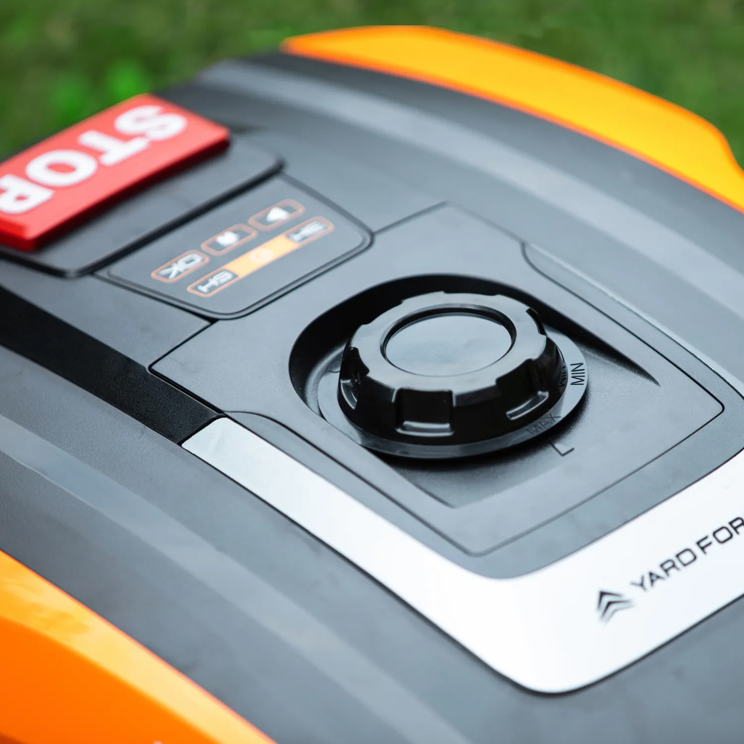 Nahaufnahme eines Mähroboters mit rotem Stoppknopf, schwarzen Bedienelementen und orangefarbenen Akzenten.