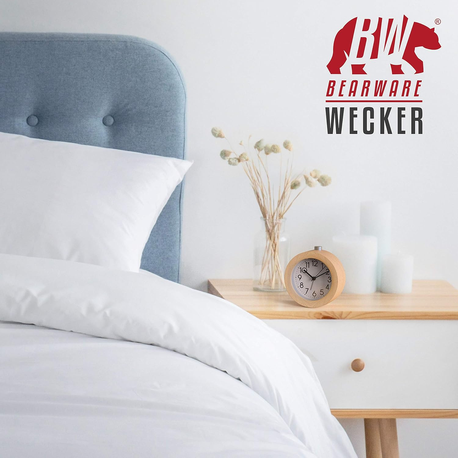 Ein Nachttisch mit einem Holzwecker, Kerzen und Trockenblumen neben einem weißen Bett.