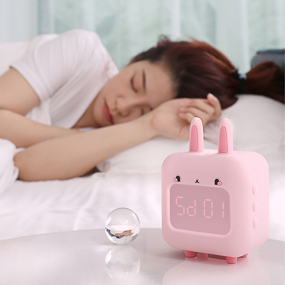 Eine Frau schläft, ein rosa Hase-Wecker und eine Glaskugel auf einem Tisch.