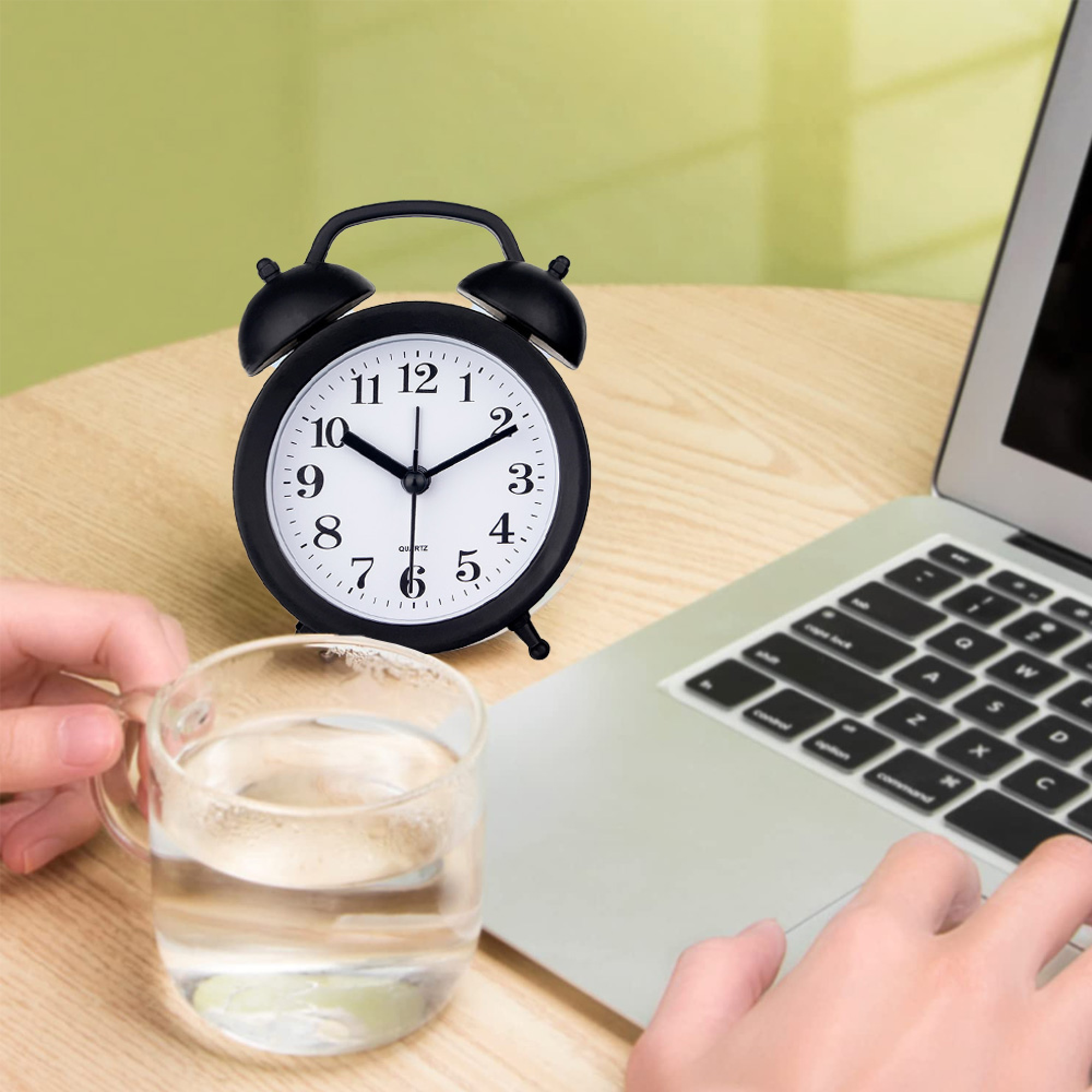 Ein schwarzer Wecker steht neben einem Glas Wasser und einem Laptop.