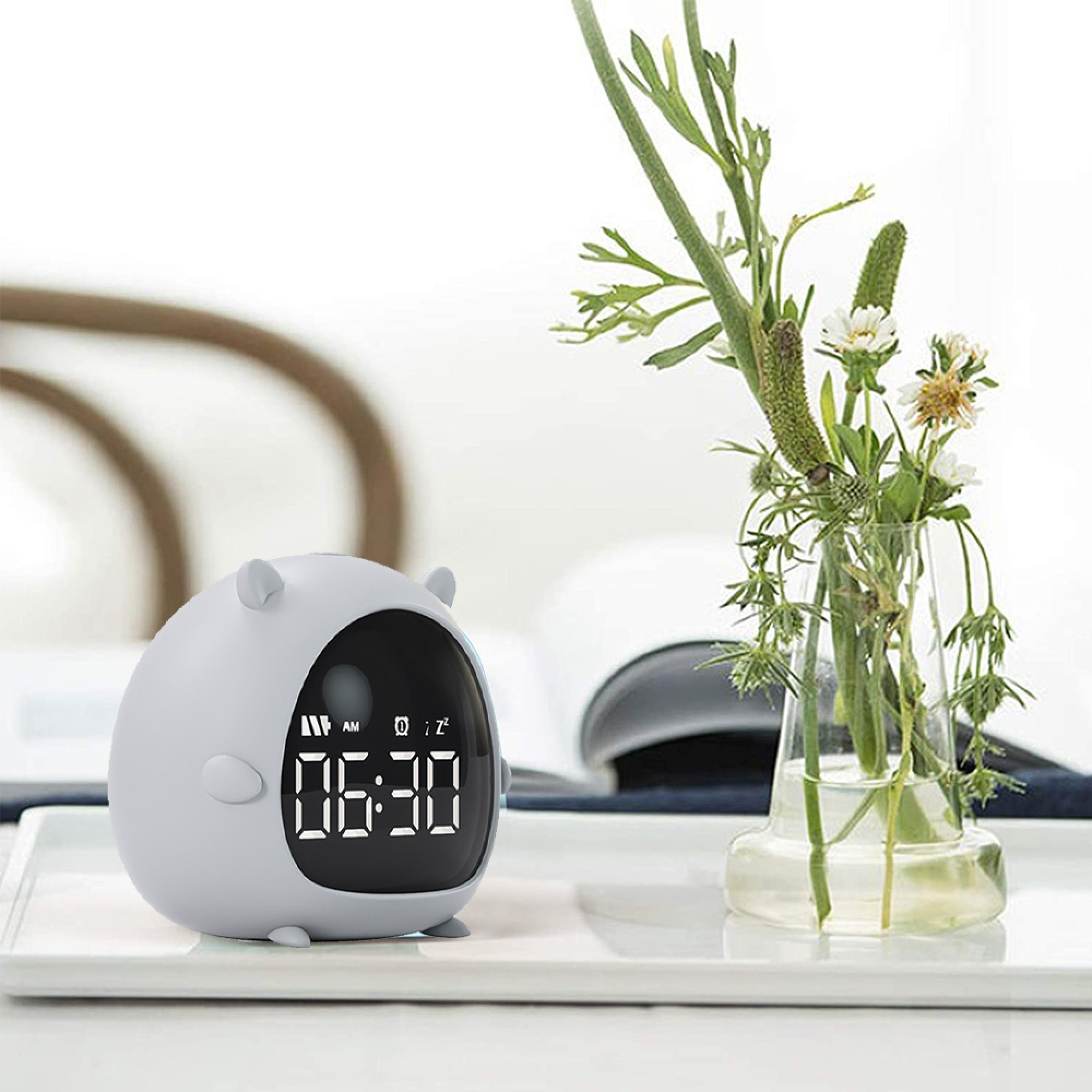 Ein süßer grauer Wecker mit Digitalanzeige, der 06:30 anzeigt, neben einer Blumenvase.