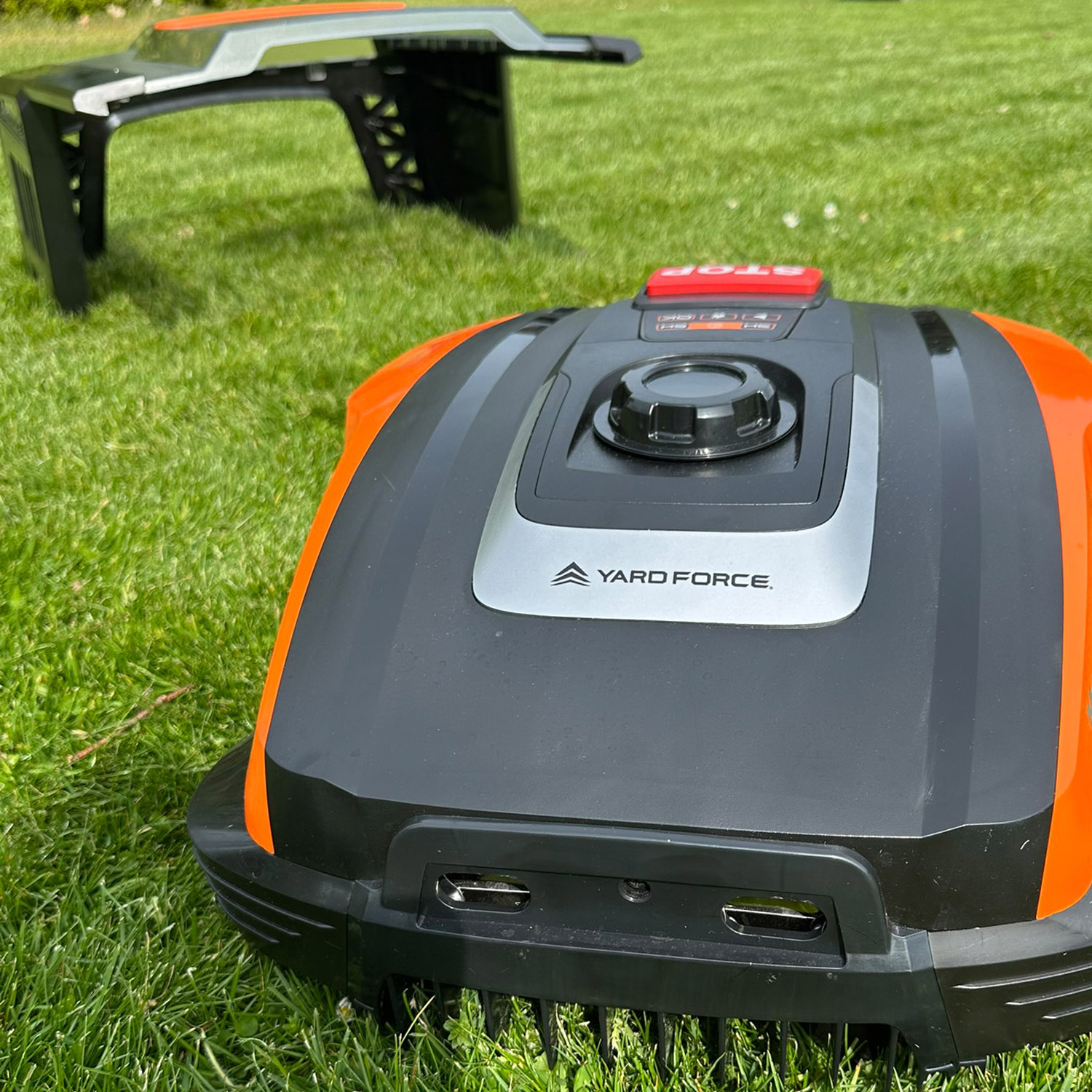 Ein orange-schwarzer Rasenmähroboter steht auf einem grünen Rasen, ein Teil eines anderen Geräts im Hintergrund.