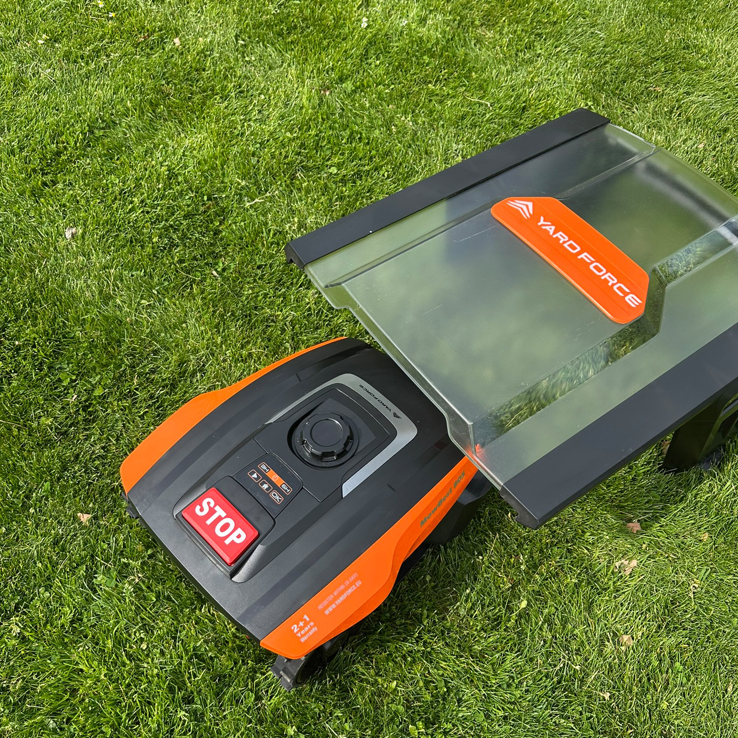 Ein Rasenmähroboter mit einer transparenten Abdeckung und orangefarbener Aufschrift liegt auf einem grünen Rasen.