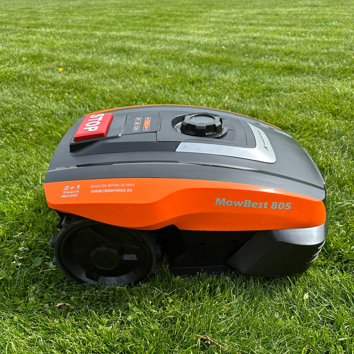 Ein orange-schwarzer Mähroboter mit der Aufschrift "MowBest 805" steht auf einem grünen Rasen.