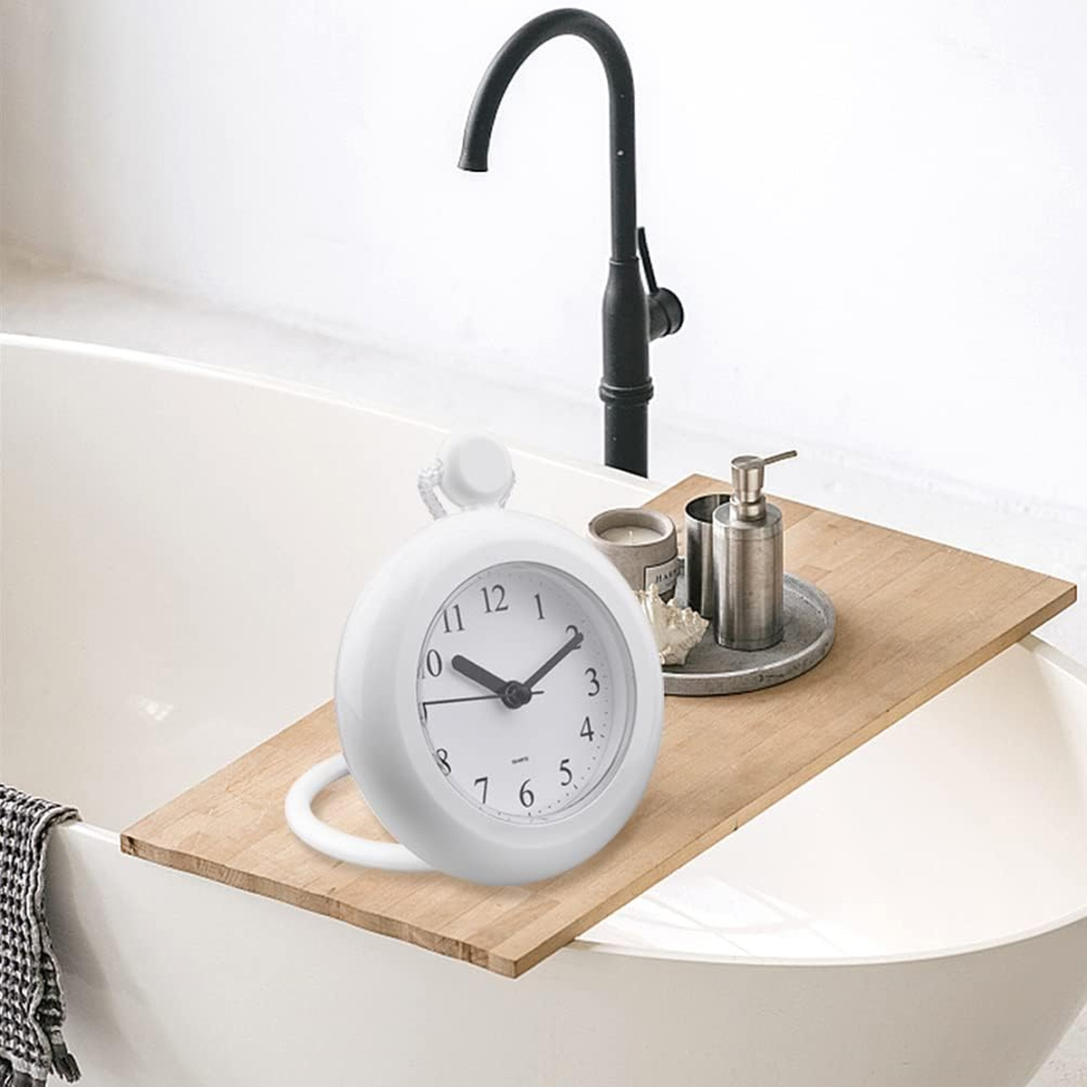 Ein weißer Badezimmerwecker auf einem Holztablett neben einer Badewanne mit schwarzem Wasserhahn.