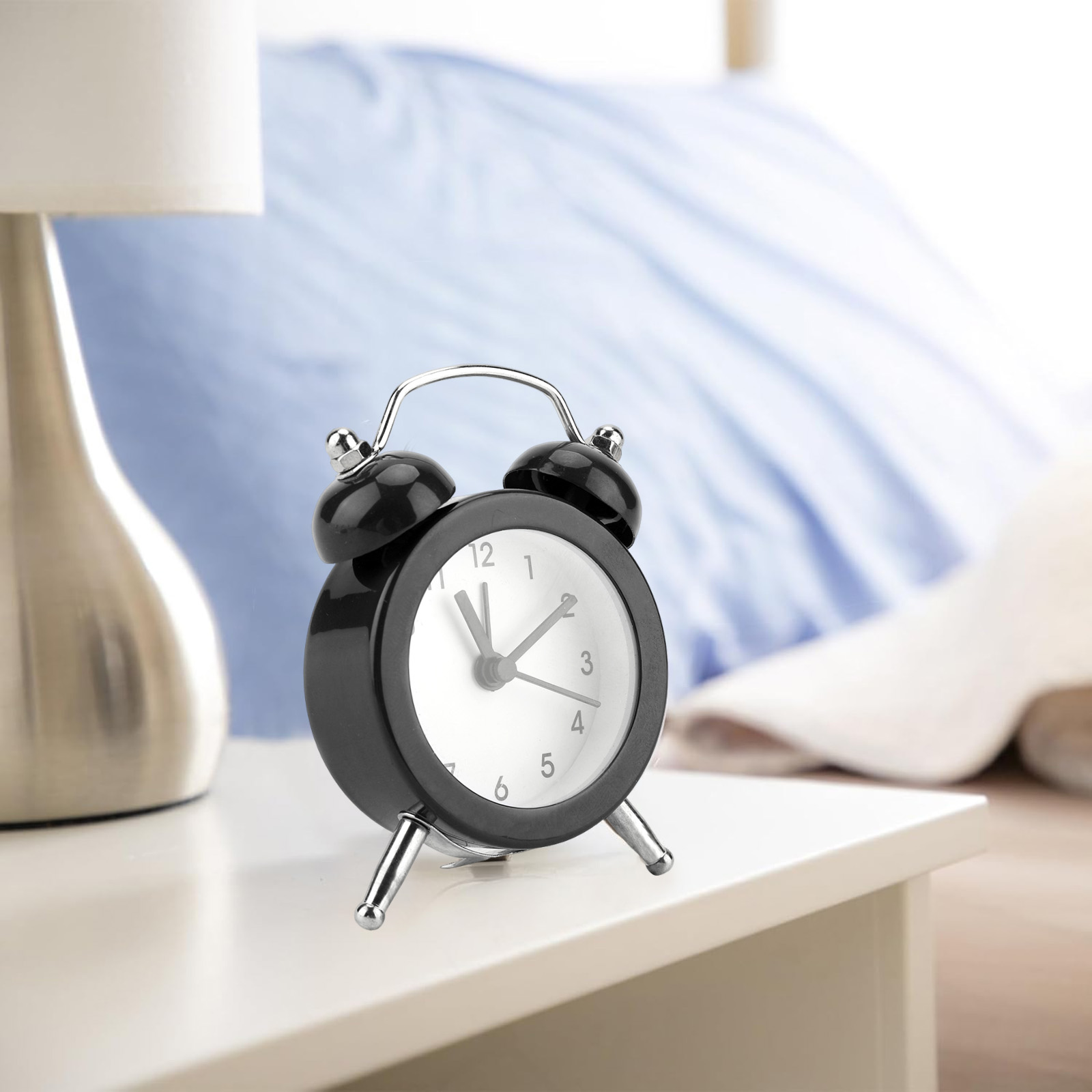 Schwarzer Wecker auf Nachttisch neben Lampe und Bett mit blauer Decke.
