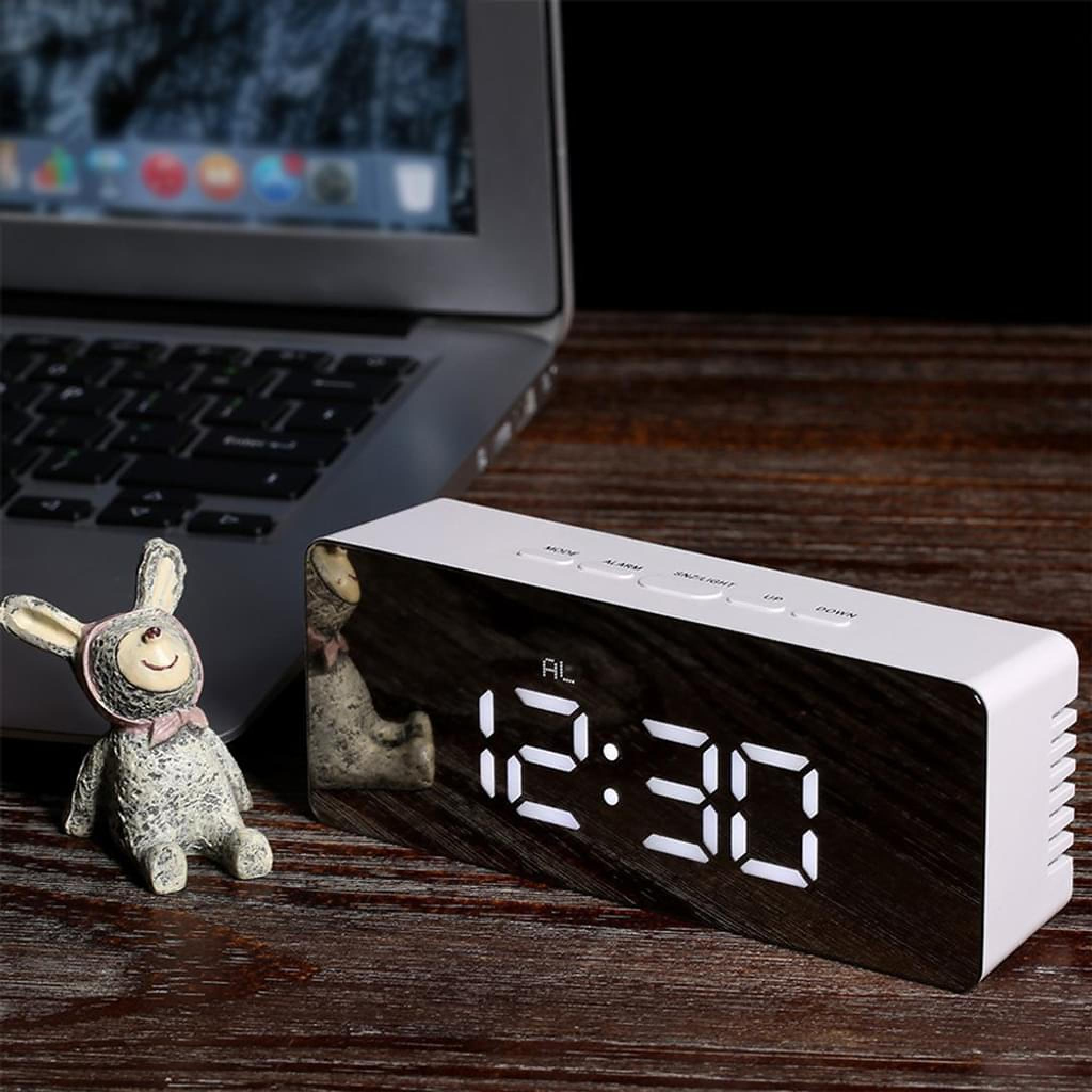 Ein digitaler Wecker mit weißen Zahlen zeigt 12:30 auf einem Holztisch neben einem Laptop und einer Hasenfigur.