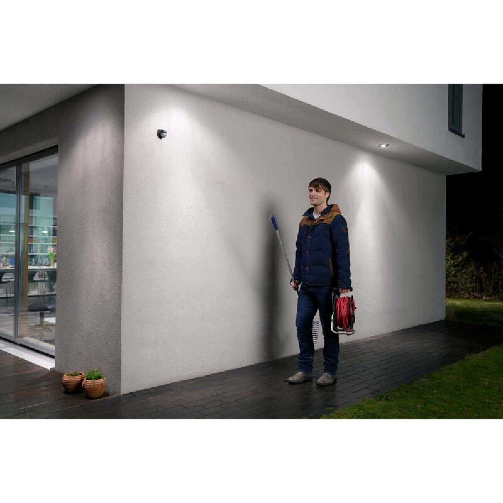 Mann in dunkler Jacke mit Kabelrolle und Stange steht auf gepflastertem Weg neben Haus.