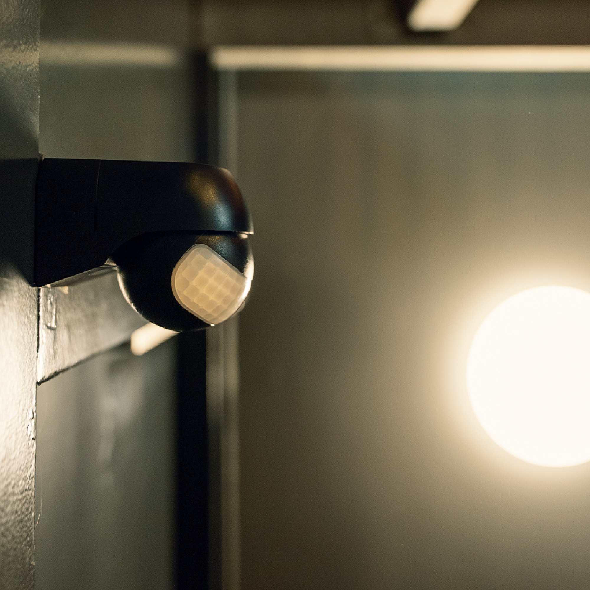 Ein schwarzer Bewegungsmelder mit einem weißen, karierten Sensor auf einer dunklen Wand.