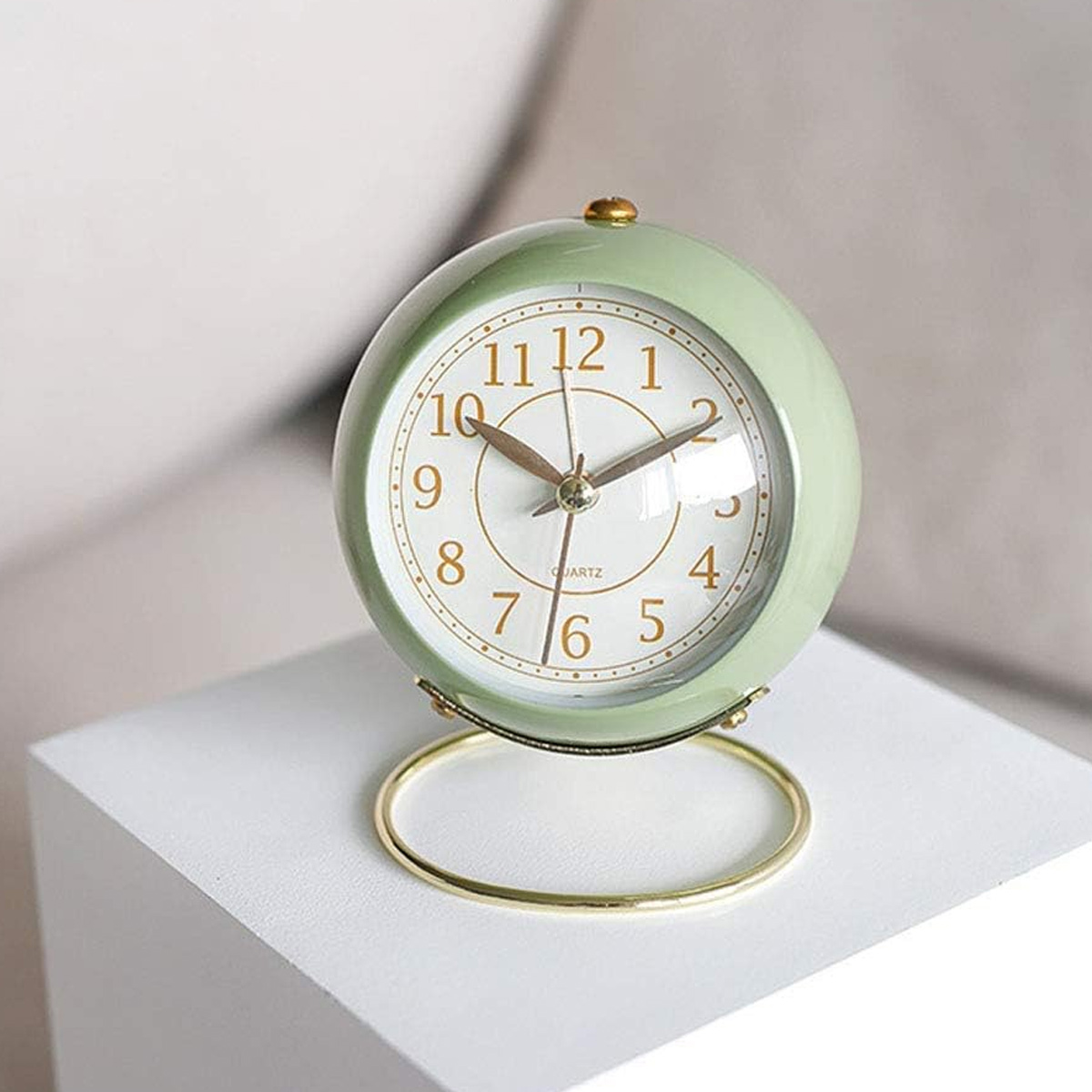 Eine hellgrüne, runde Weckuhr mit goldenem Ständer steht auf einer weißen Fläche.