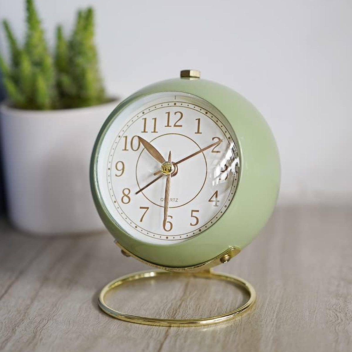 Eine hellgrüne Kugelweckeruhr auf einem goldenen Ständer mit Pflanzen im Hintergrund.