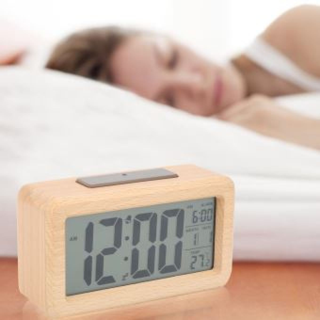 Ein hölzerner Digitalwecker zeigt 12:00 an, eine Person schläft im Hintergrund.
