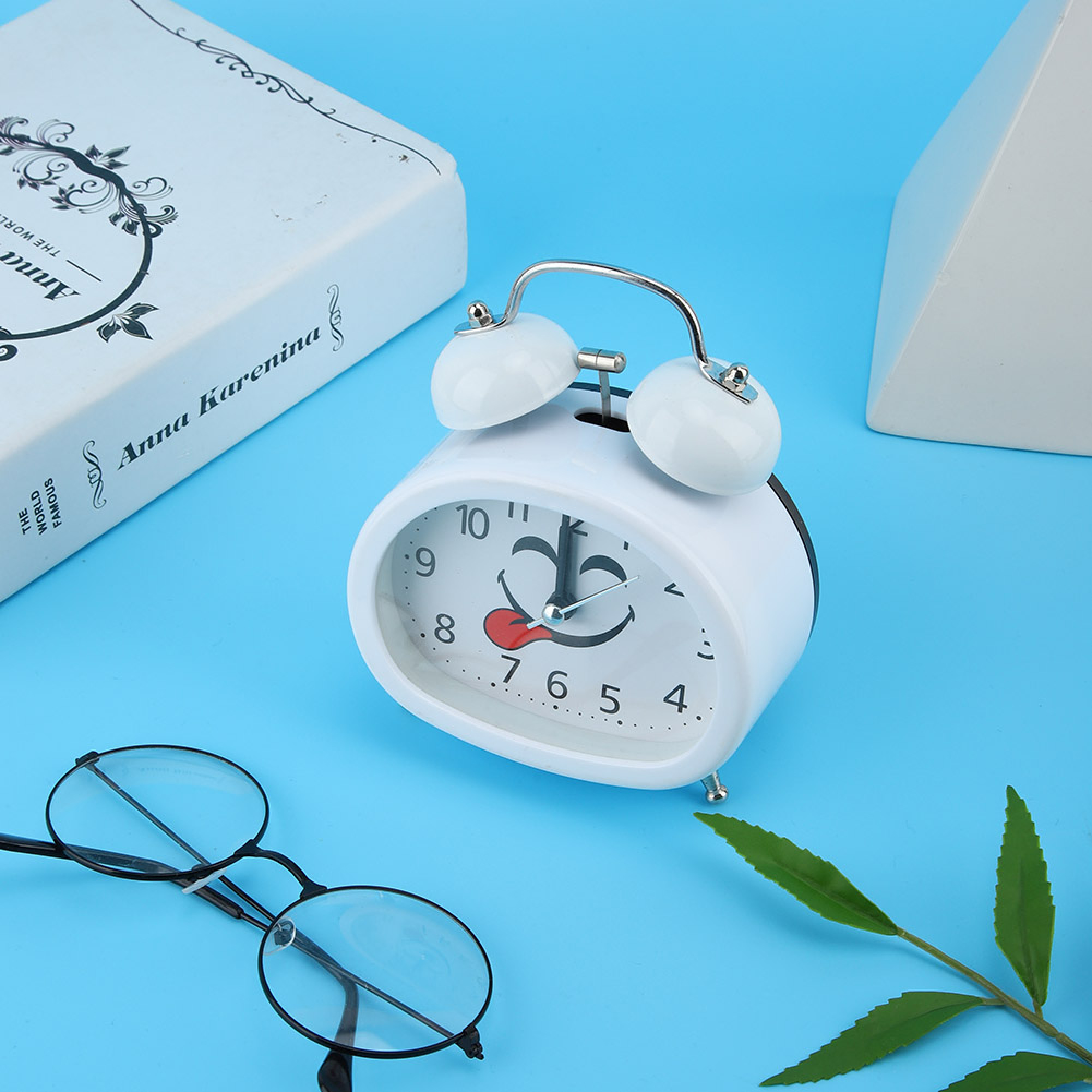 Ein weißer Wecker mit Smiley-Gesicht neben einem Buch und Brille.