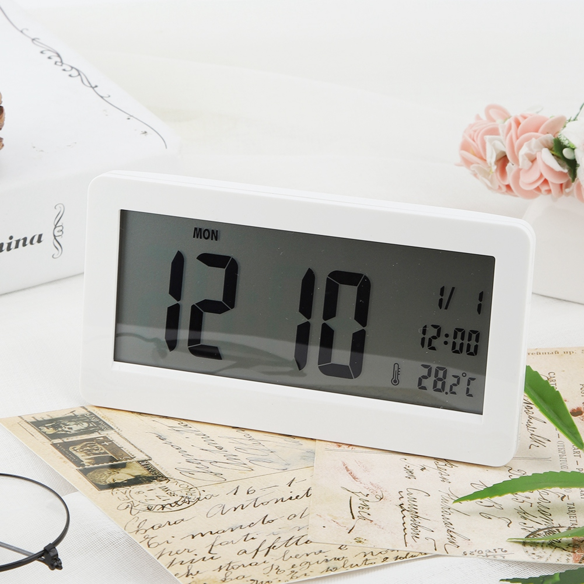 Eine weiße Digitaluhr zeigt MON 12:10, 1/1, 12:00 und 28,2C auf Vintage-Postkarten.