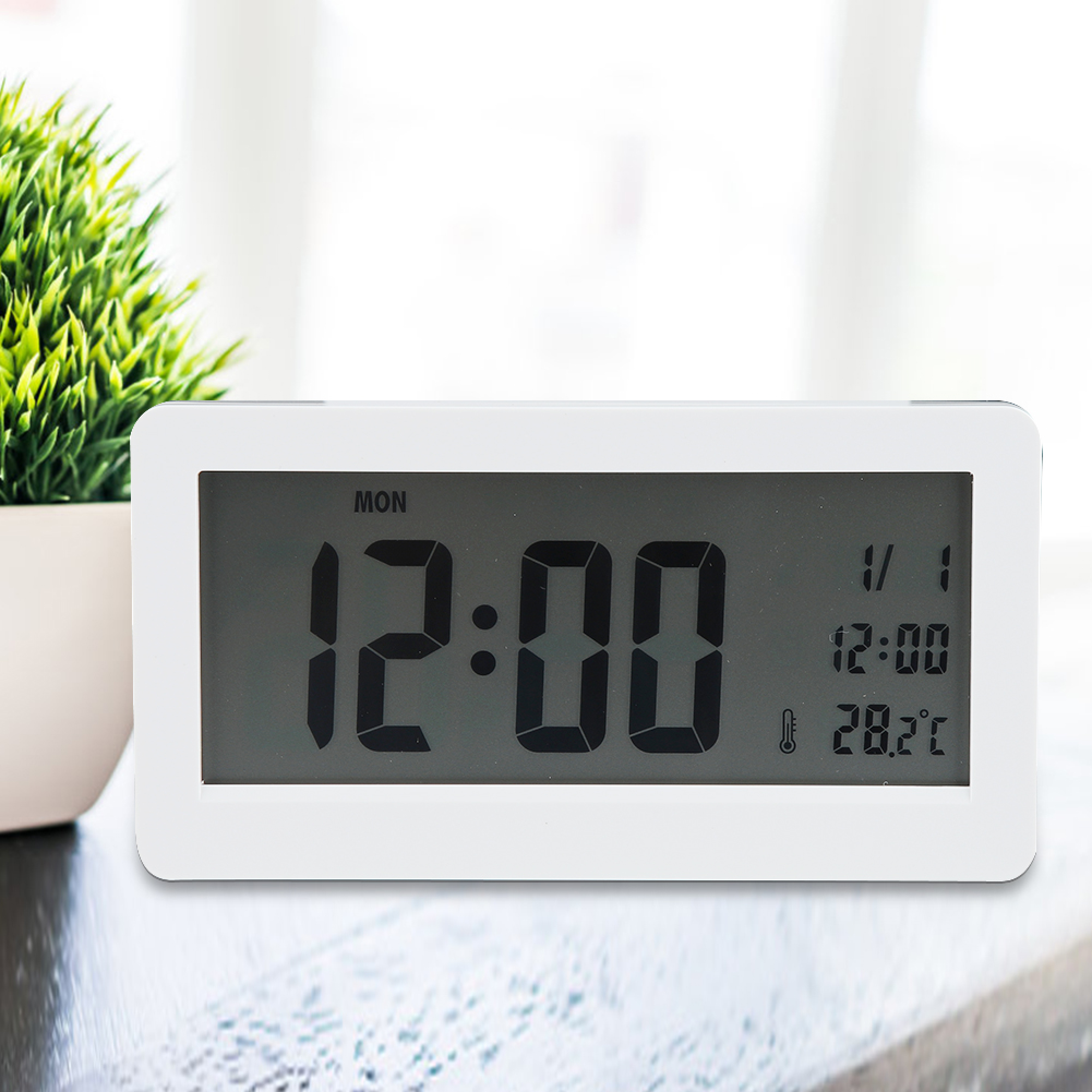 Ein weißer Digitalwecker zeigt 12:00 Uhr und 28,2°C an, neben einer grünen Topfpflanze.