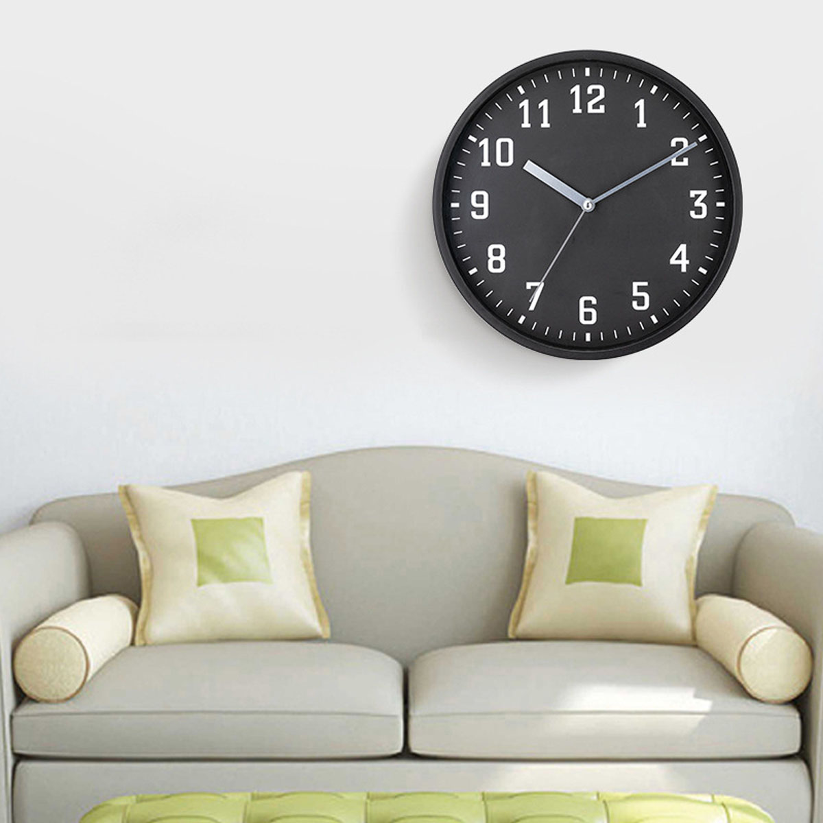 Eine schwarze Wanduhr mit weißen Zahlen hängt über einem neutralen Sofa mit Zierkissen.