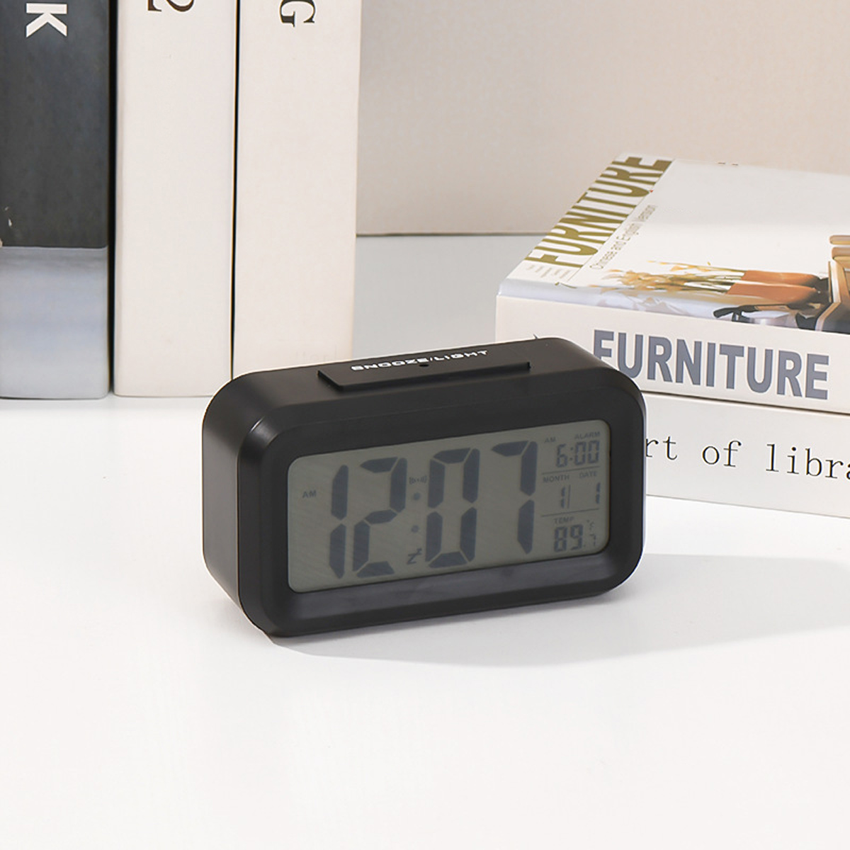 Ein schwarzer Digitalwecker zeigt 12:07 Uhr an. Im Hintergrund sind Bücher.