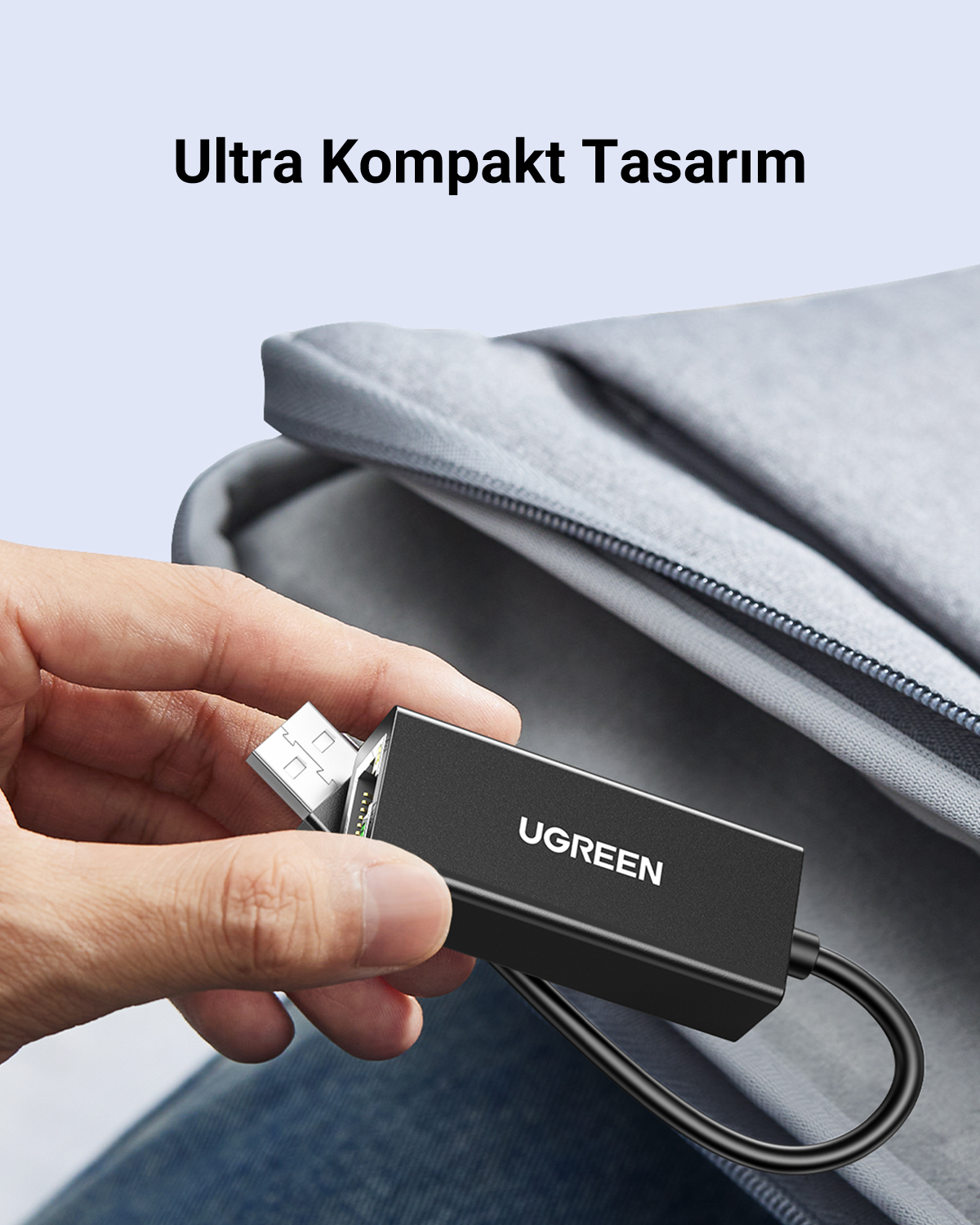 Bir el, gri bir çantanın yanında siyah bir UGREEN USB - Ethernet adaptörü tutuyor.