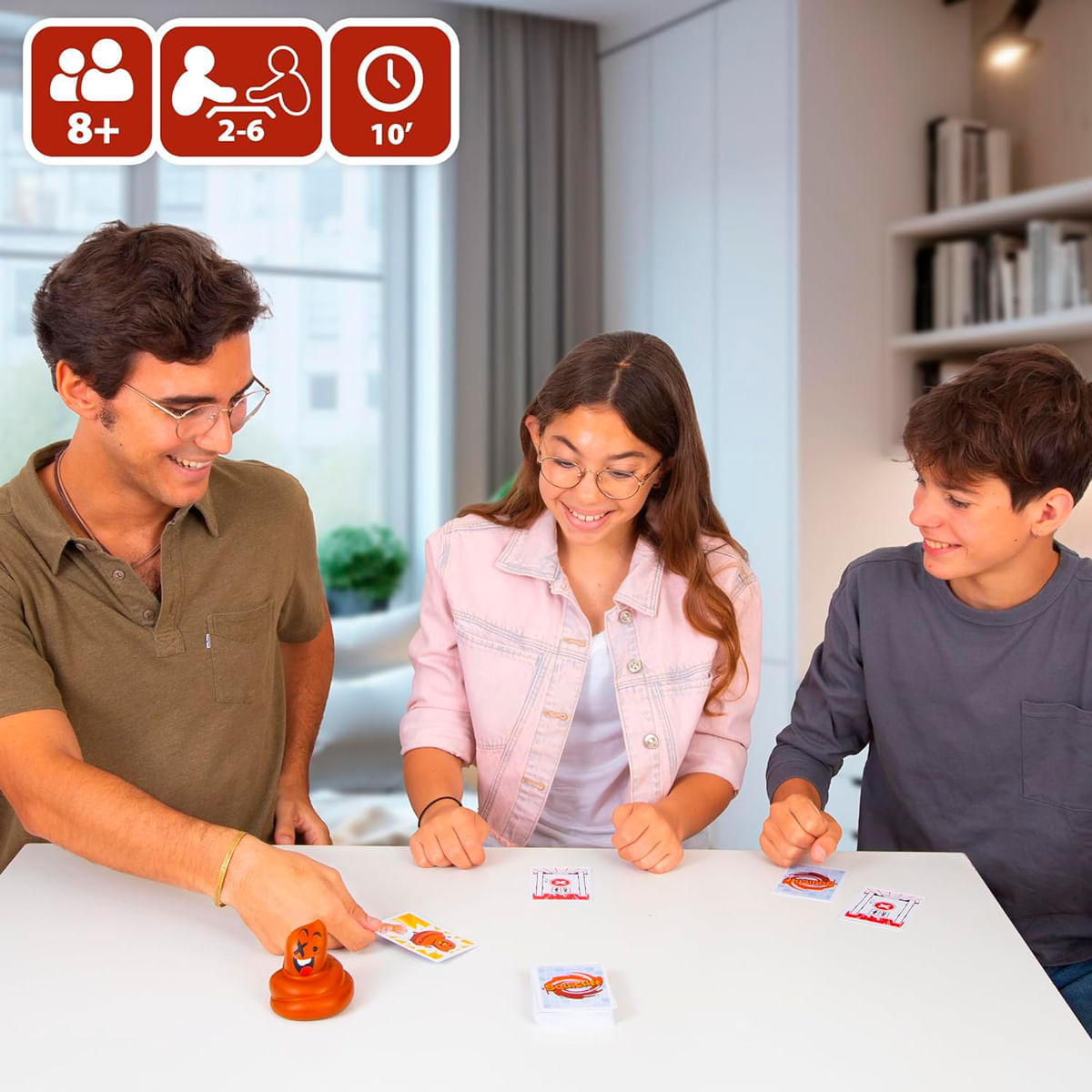 Tres amigos sonrientes juegan a un juego de cartas con una figura de perro marrón sobre una mesa blanca.