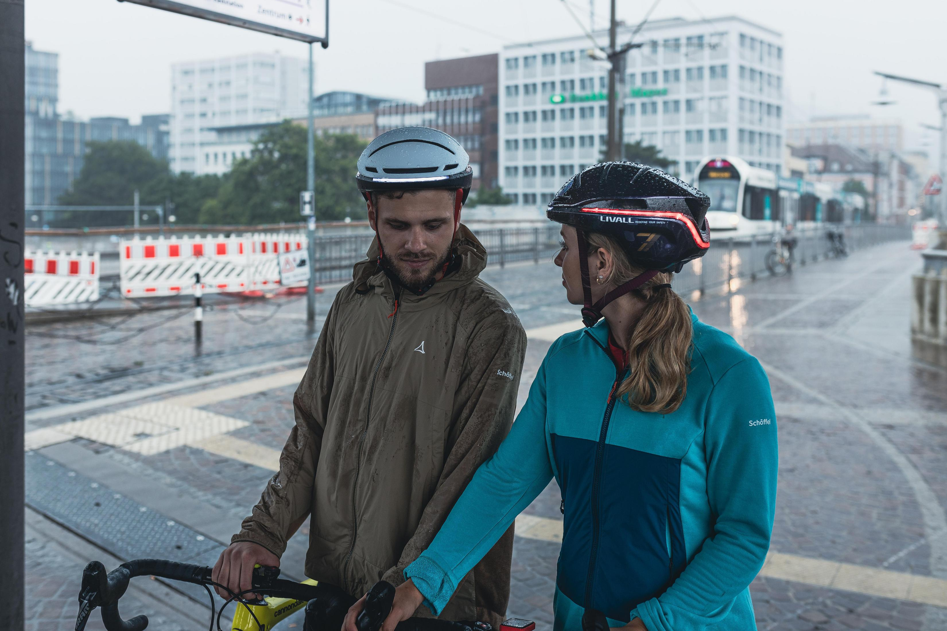 Due ciclisti con casco sotto la pioggia. Un tram sullo sfondo.