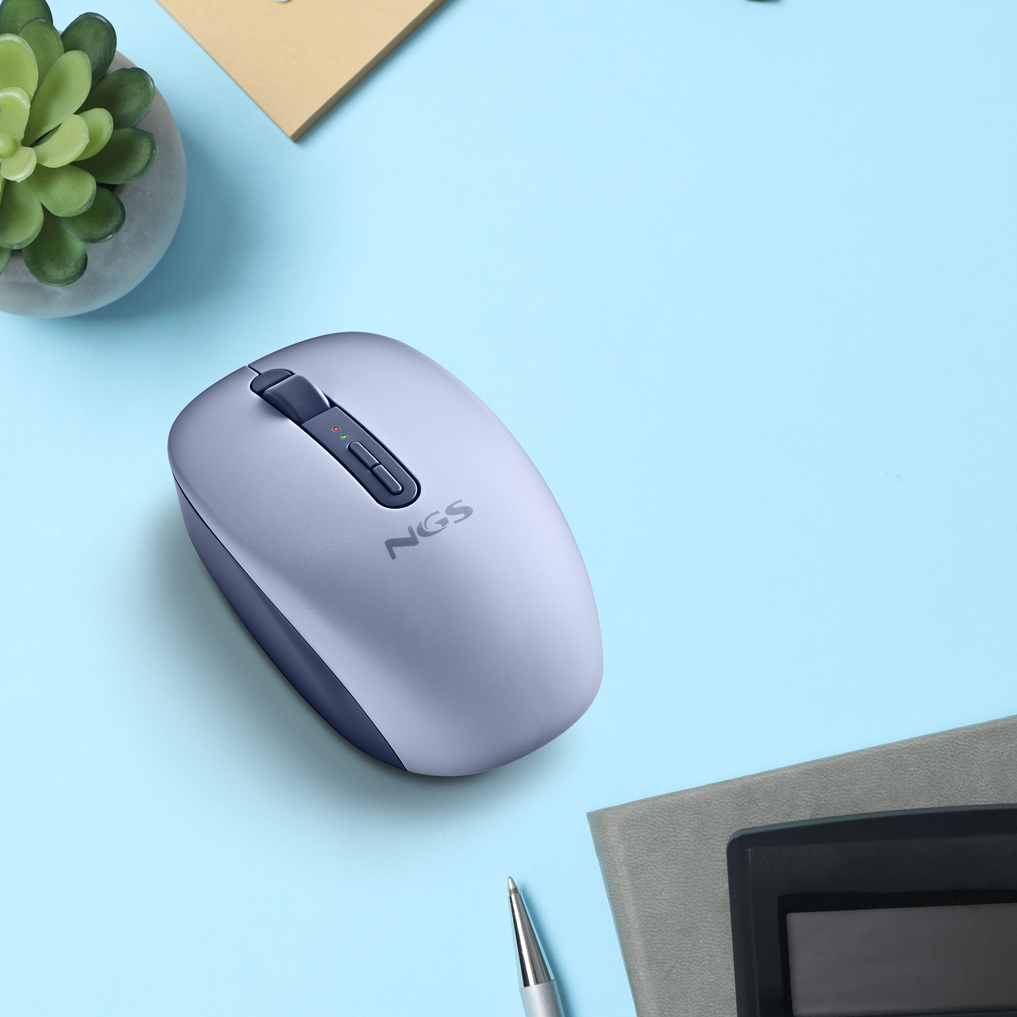 Mouse NGS grigio su scrivania blu con pianta, taccuino e penna.