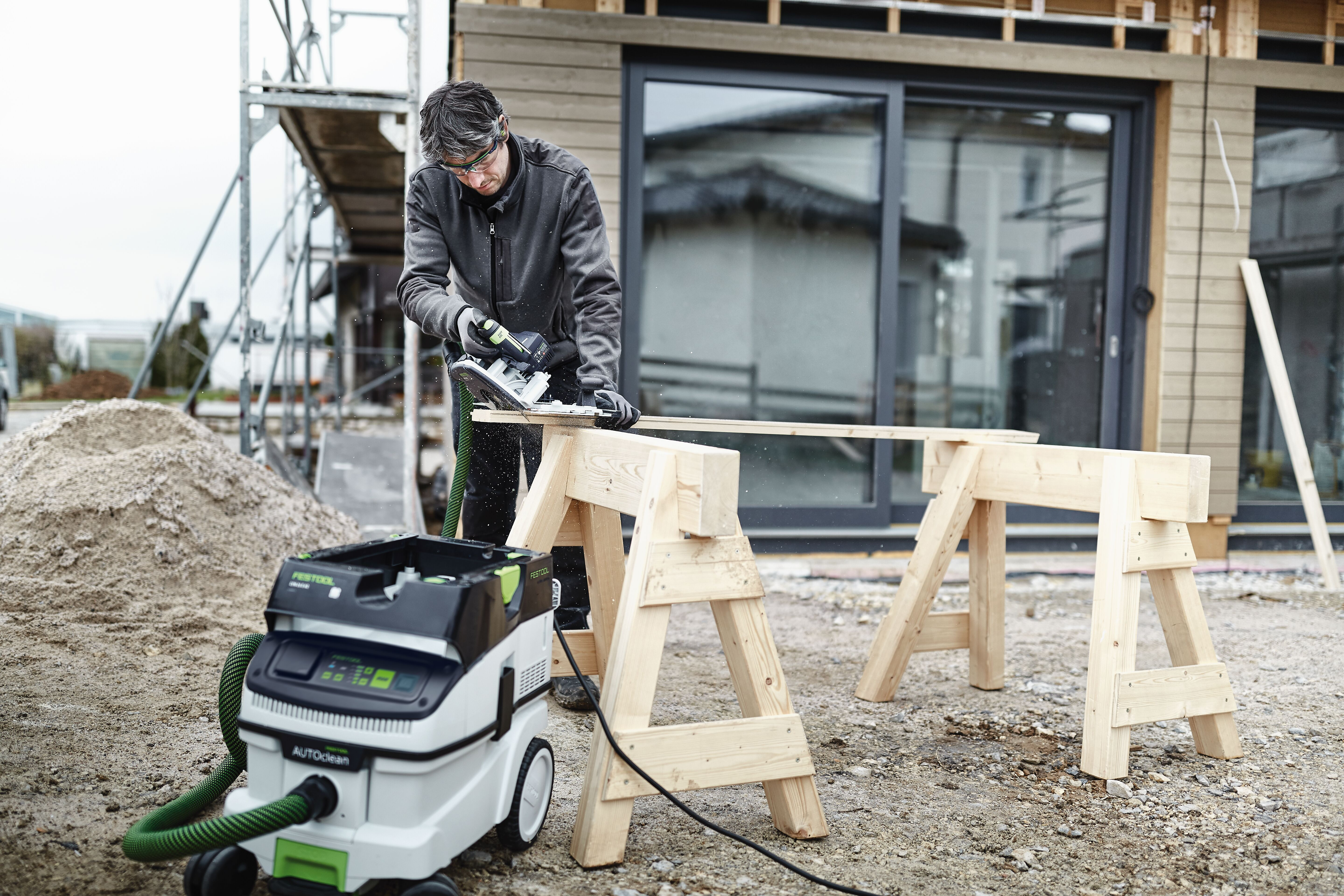 Ein Mann sägt Holz auf Baustelle, daneben ein Staubsauger und Sandhaufen.