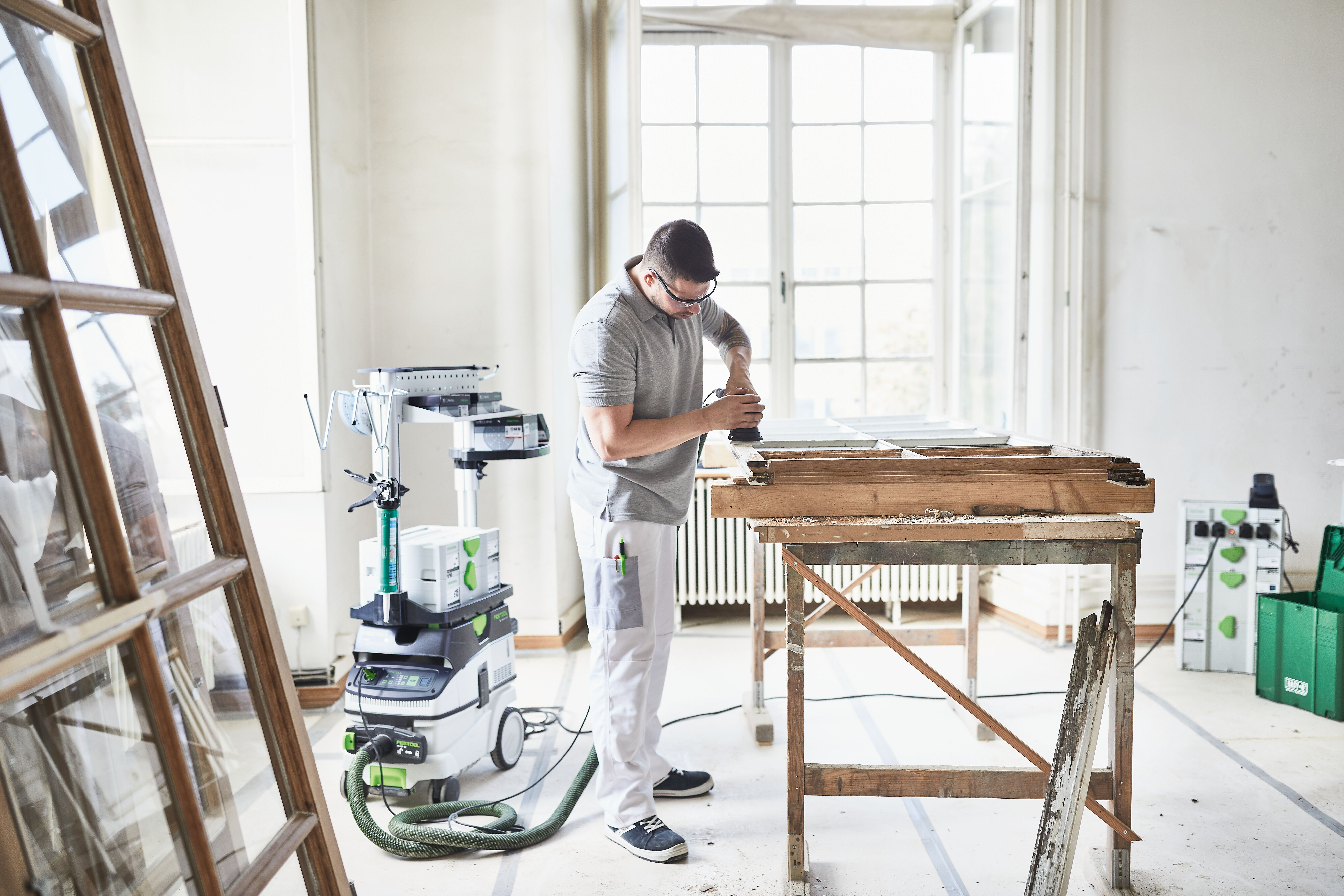 Mann schleift Holz auf Werkbank, Staubsauger und Leiter daneben.