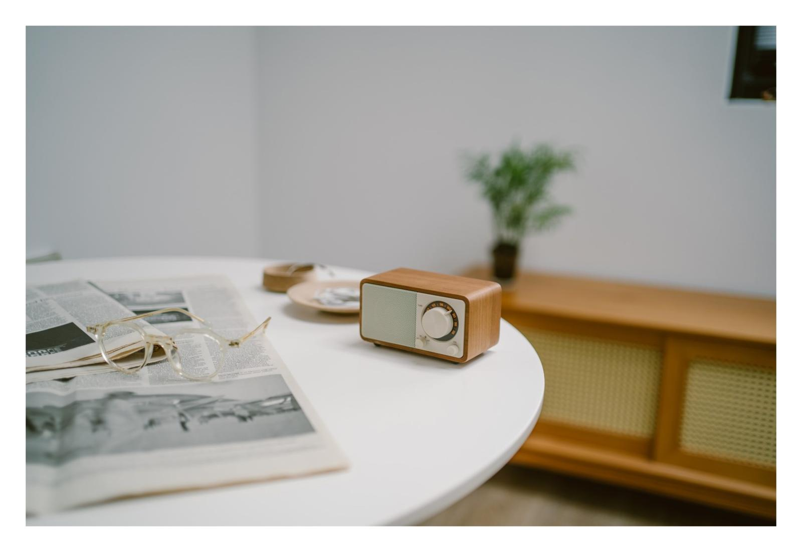 Ein Holzradio steht auf einem weißen Tisch neben einer Zeitung und einer Brille.