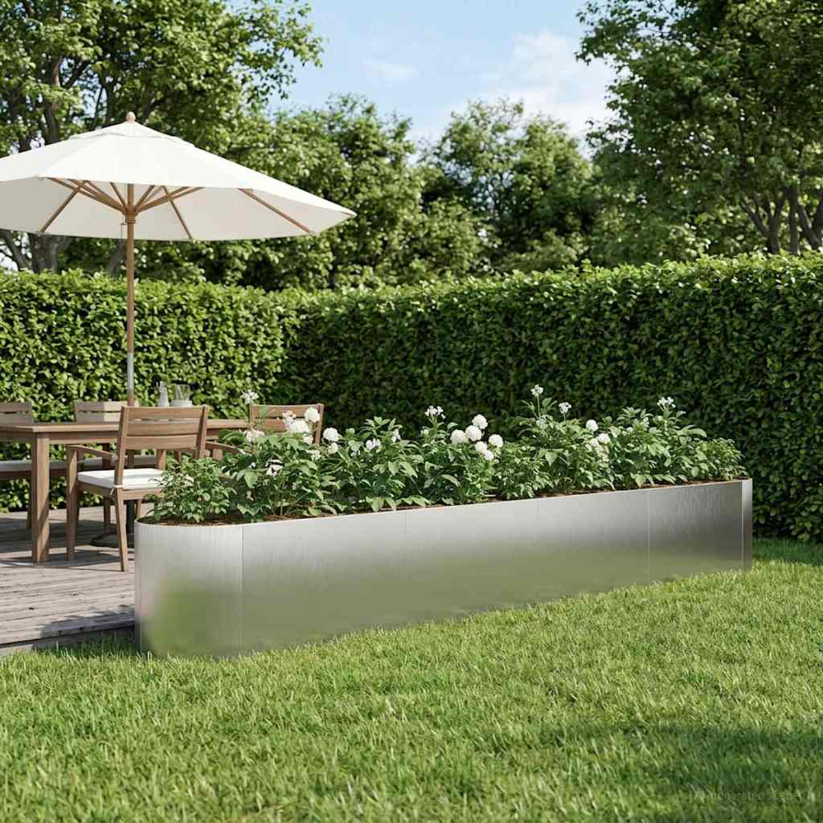Ein Metallpflanzer mit weißen Blumen steht auf Gras, nahe einer Holzterrasse mit Stühlen.