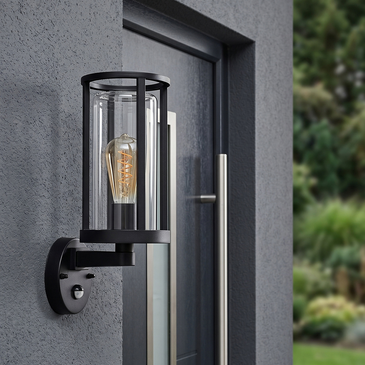 Schwarze Außenwandleuchte mit Glaszylinder und Glühbirne vor dunkler Tür.