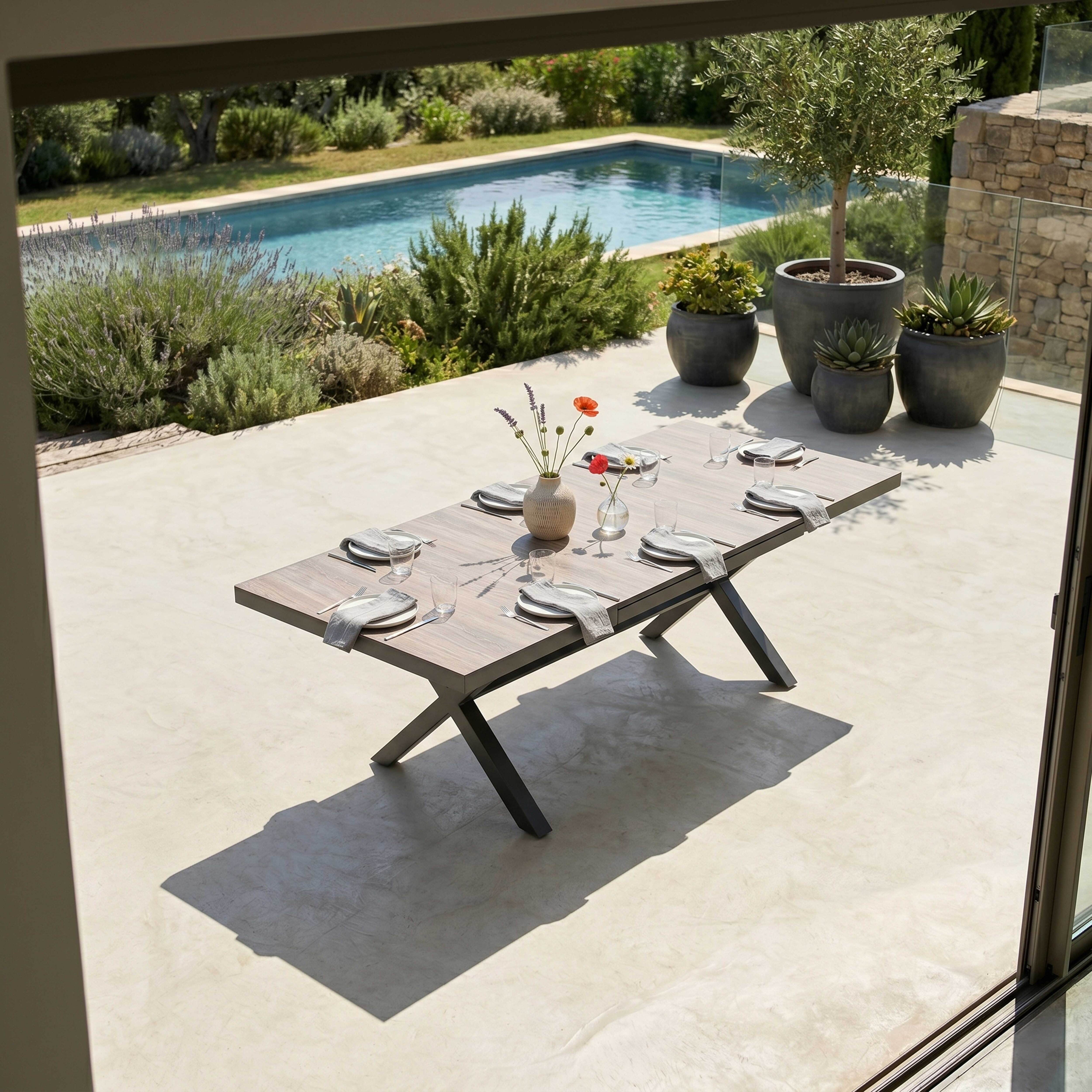 Ein Holztisch mit gedeckten Tellern, Gläsern und Blumen im Freien mit Pool und Grün.