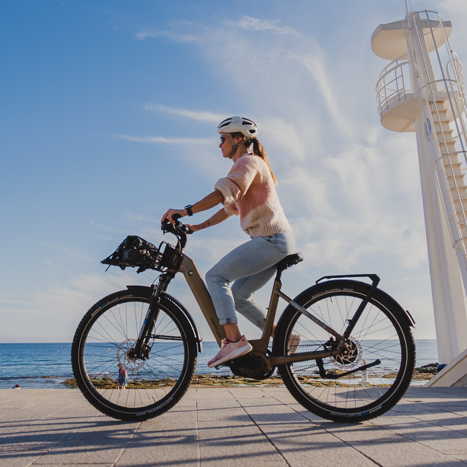 Frau fährt Fahrrad am Meer, weiße Turmstruktur im Hintergrund.