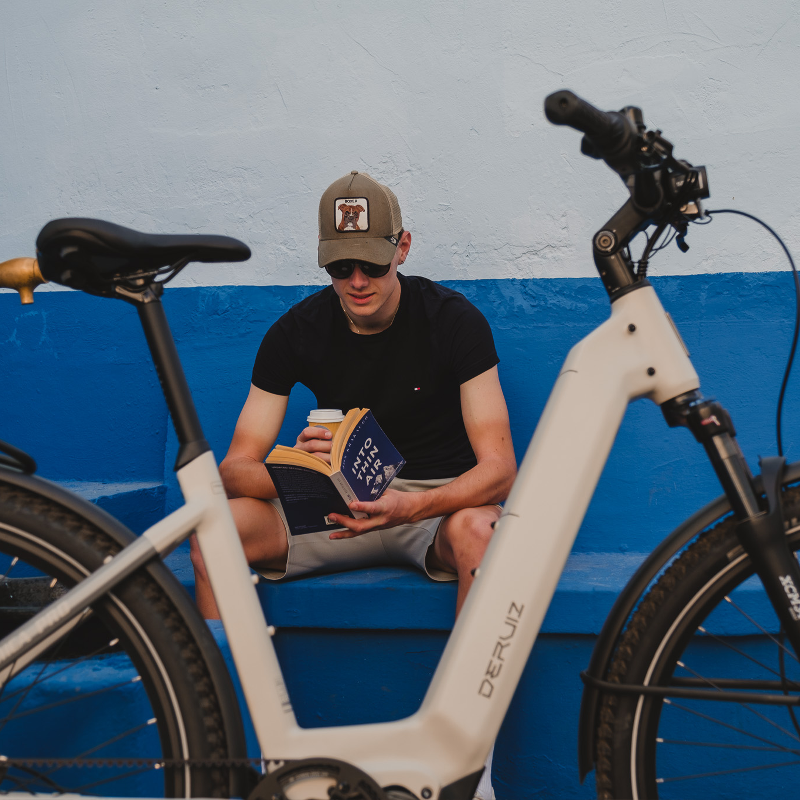 Ein Mann sitzt auf einer blauen Bank und liest ein Buch neben einem E-Bike.