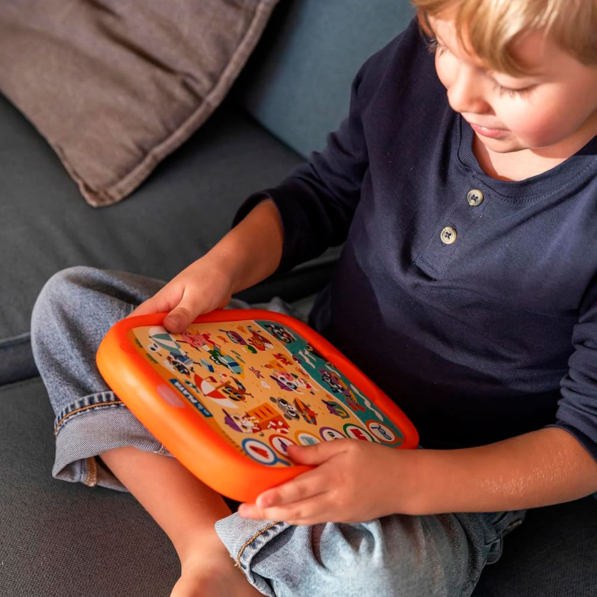 Un niño con jeans y camisa azul marino sostiene un juguete de aprendizaje naranja con imágenes coloridas.