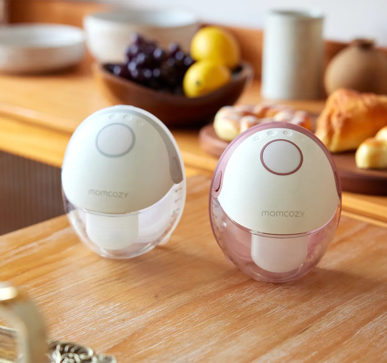 Due tiralatte Momcozy, uno bianco, uno rosa, su legno con frutta e pasticcini.